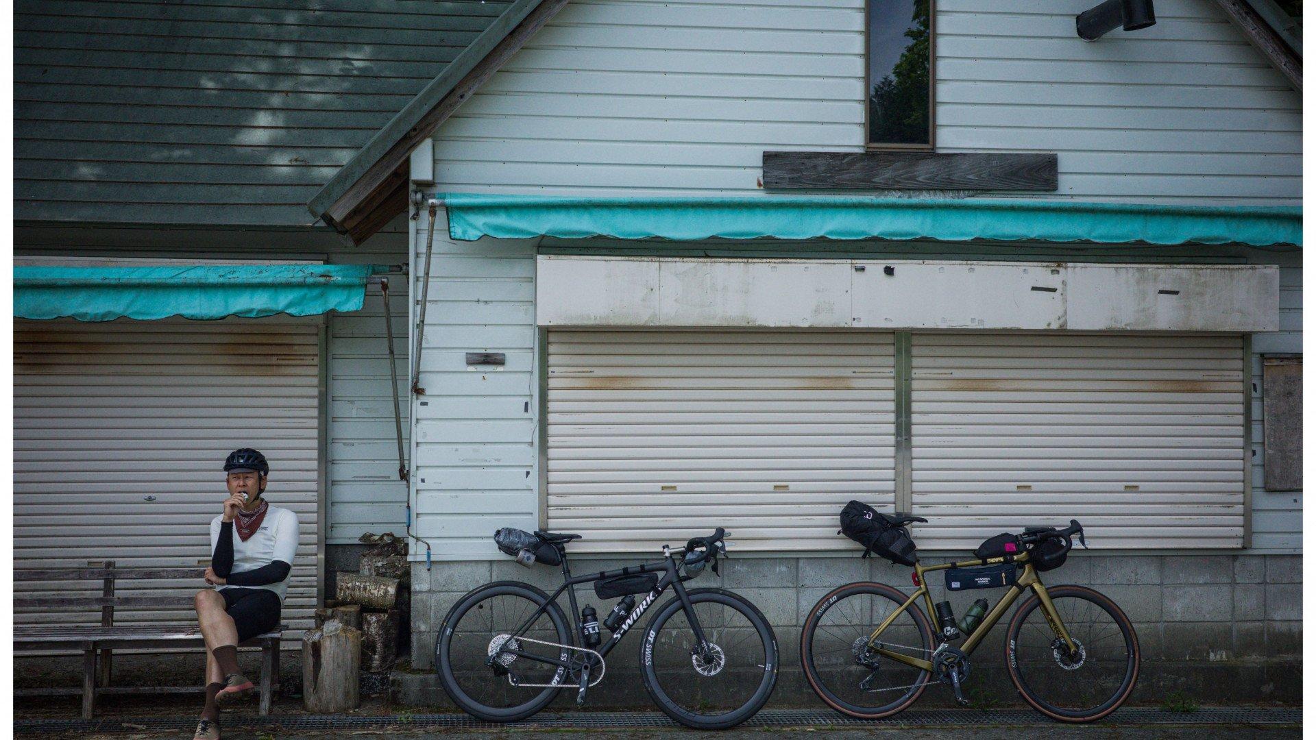 Model wearing white long-sleeve jersey and black shorts with neck warmer, sitting beside bikepacking bikes at Japanese storefront