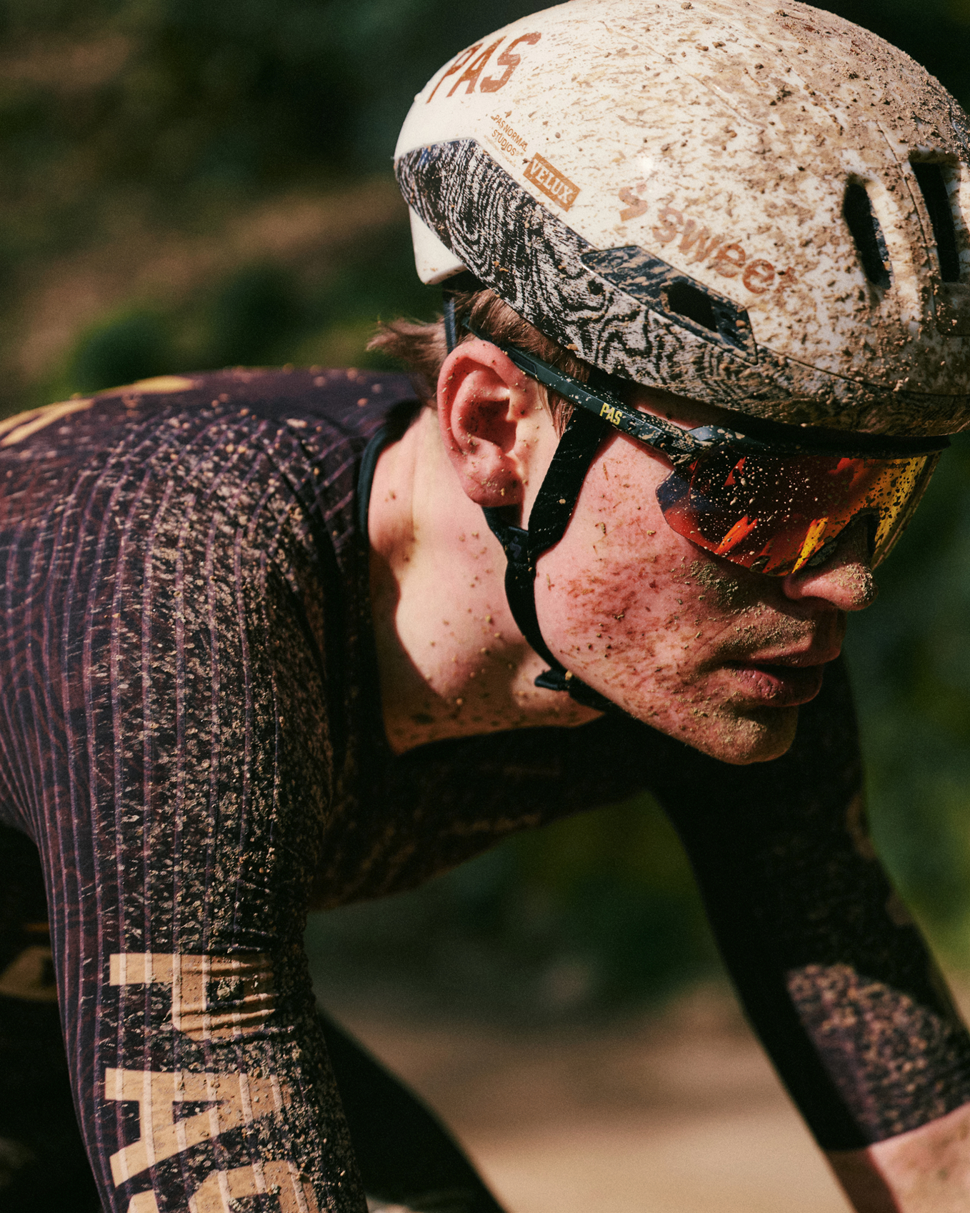 Cyclist wearing white helmet and mirrored sunglasses in aerodynamic position, mud-splattered from racing