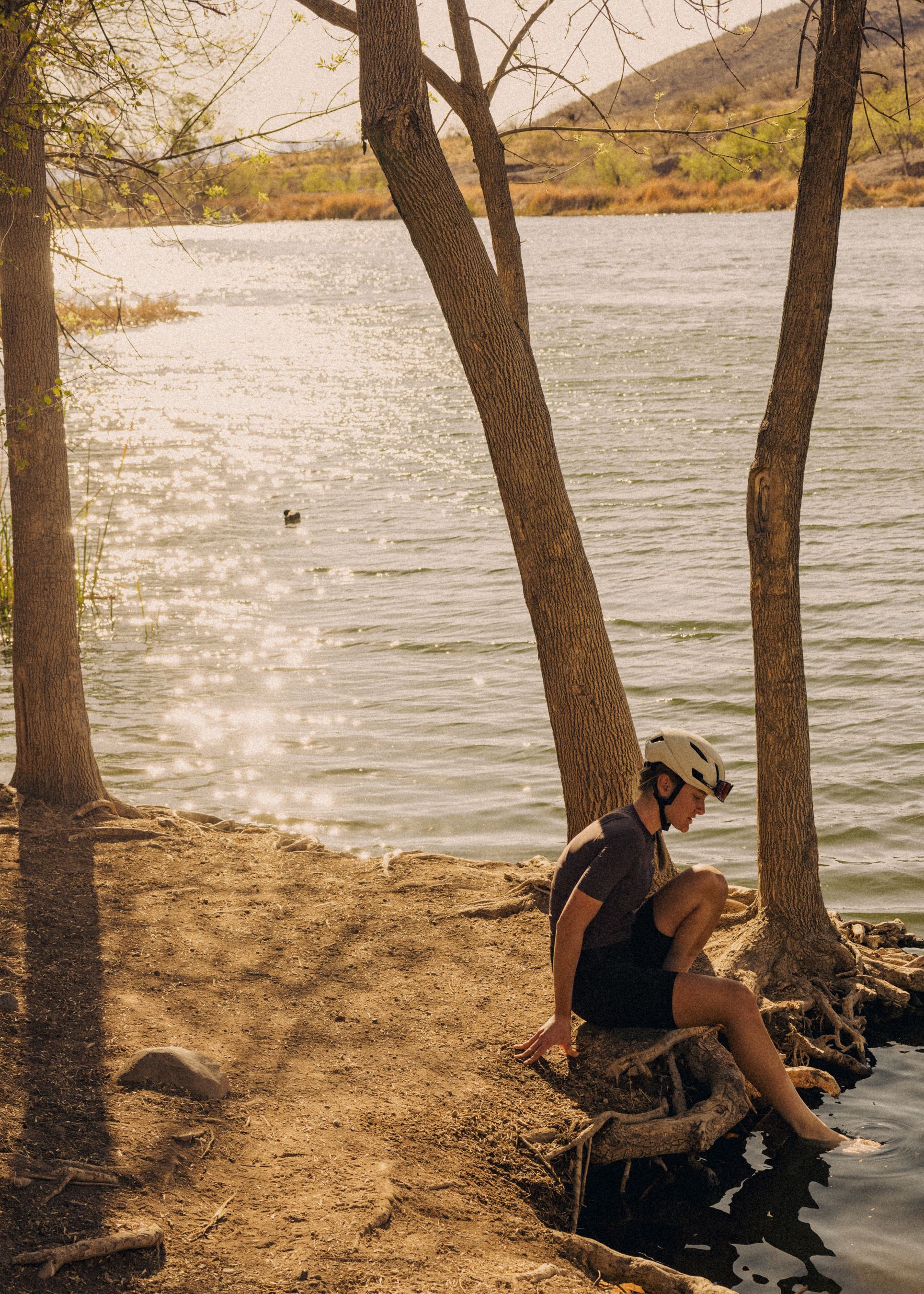 Model wearing dark cycling kit and helmet sitting by lake shore among trees