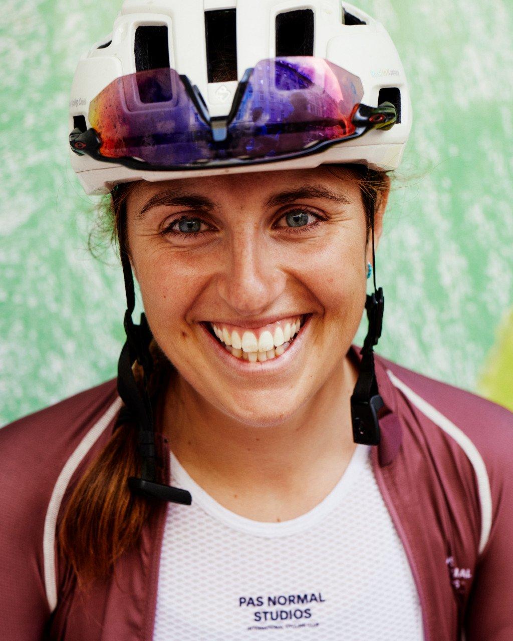 Model wearing white cycling jersey and burgundy jacket with white helmet and reflective visor, smiling outdoors