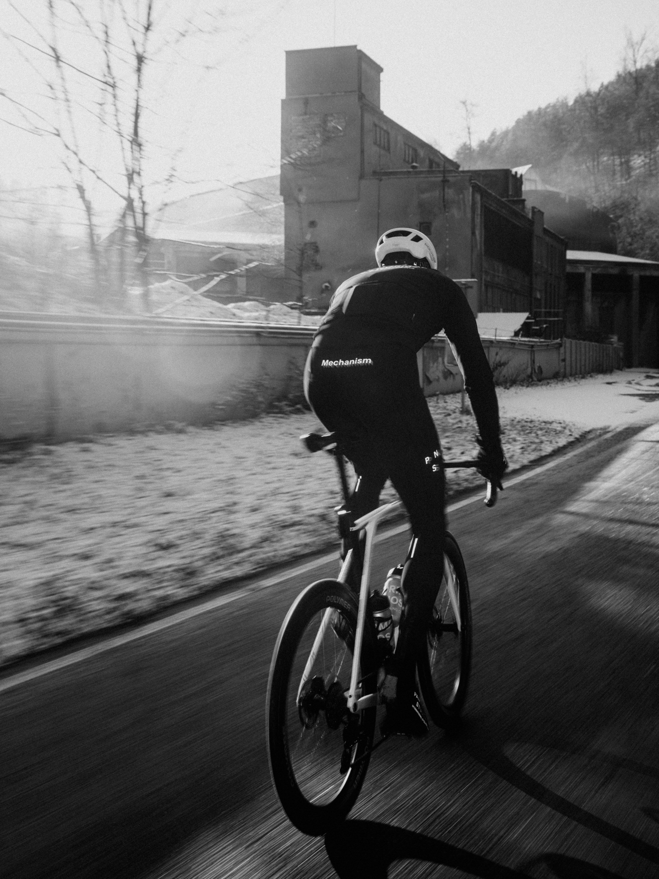 Model wearing black Mechanism cycling kit in aerodynamic position on time trial bike in industrial setting