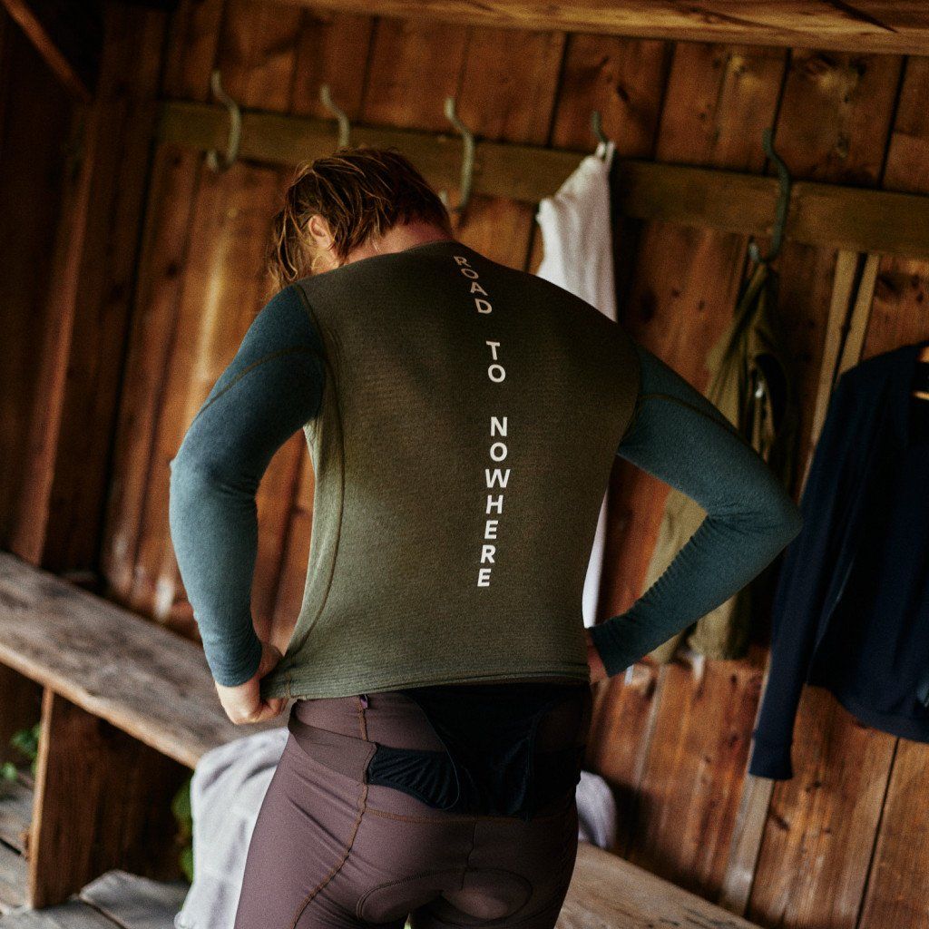 Model wearing green and blue long sleeve jersey with vertical spine text, viewed from behind in wooden cabin