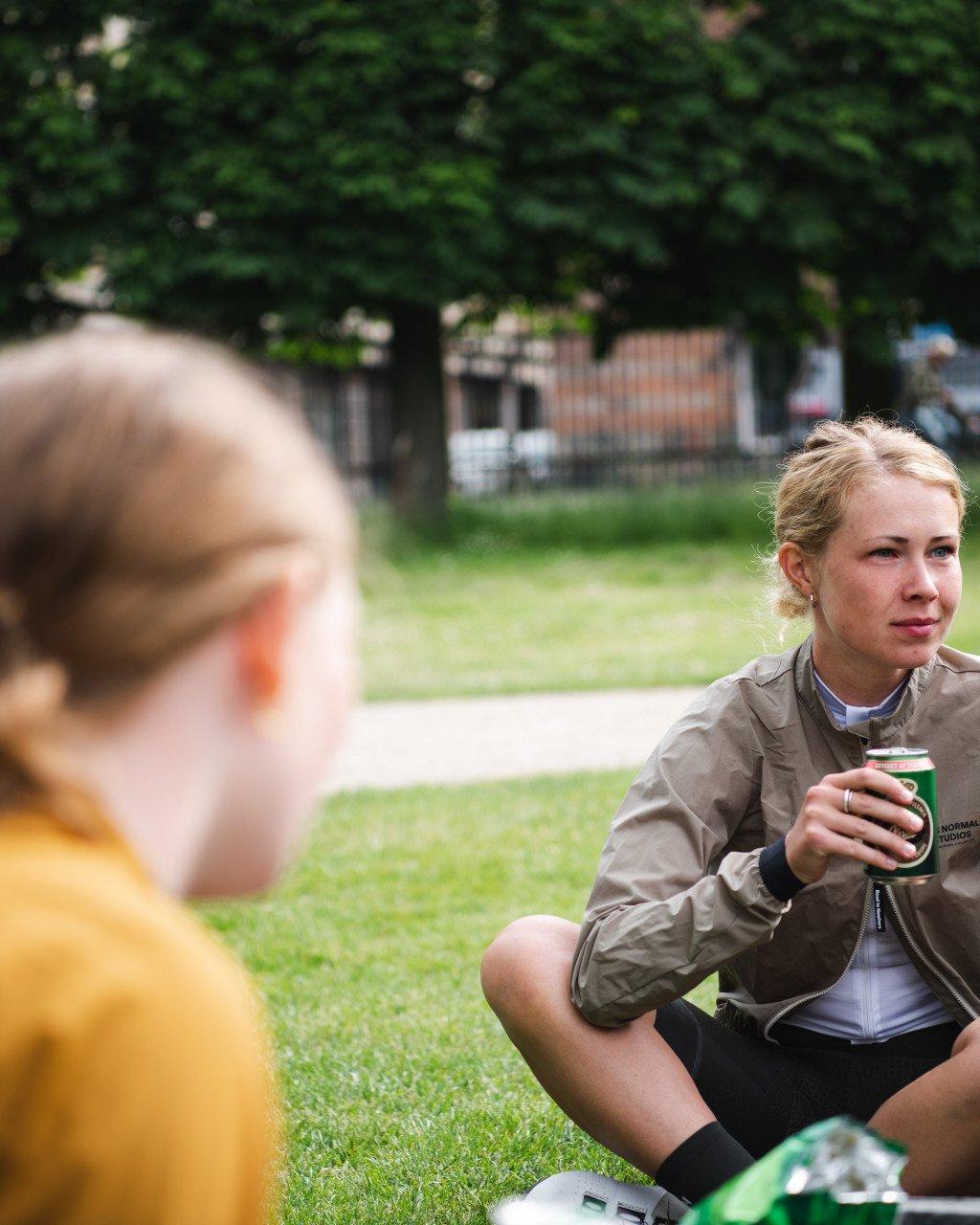 Model wearing beige cycling jacket and black shorts sitting on grass holding drink in park setting