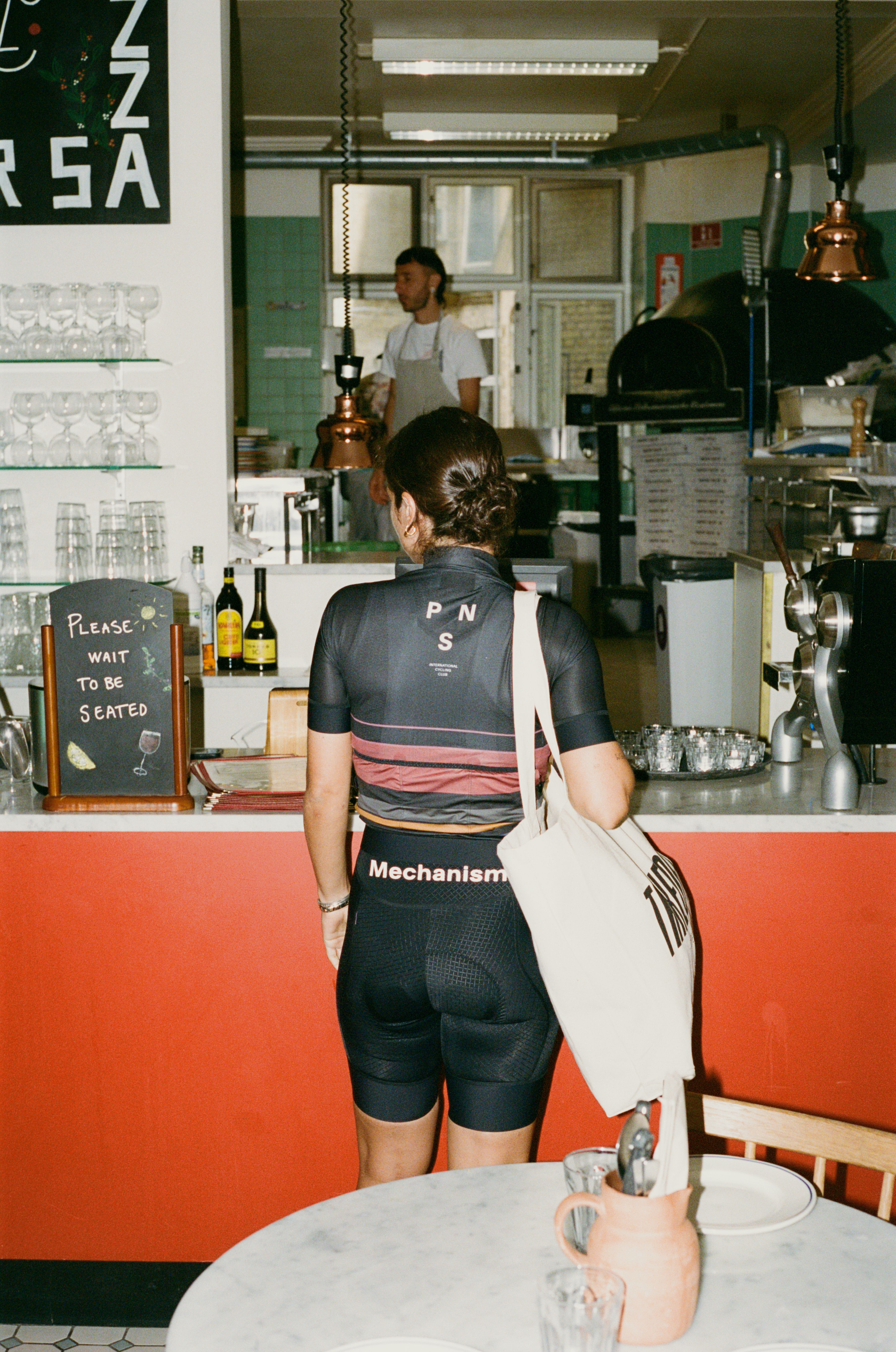 Model wearing Women's Mechanism jersey and bib shorts in dark grey with coral stripe details in restaurant kitchen