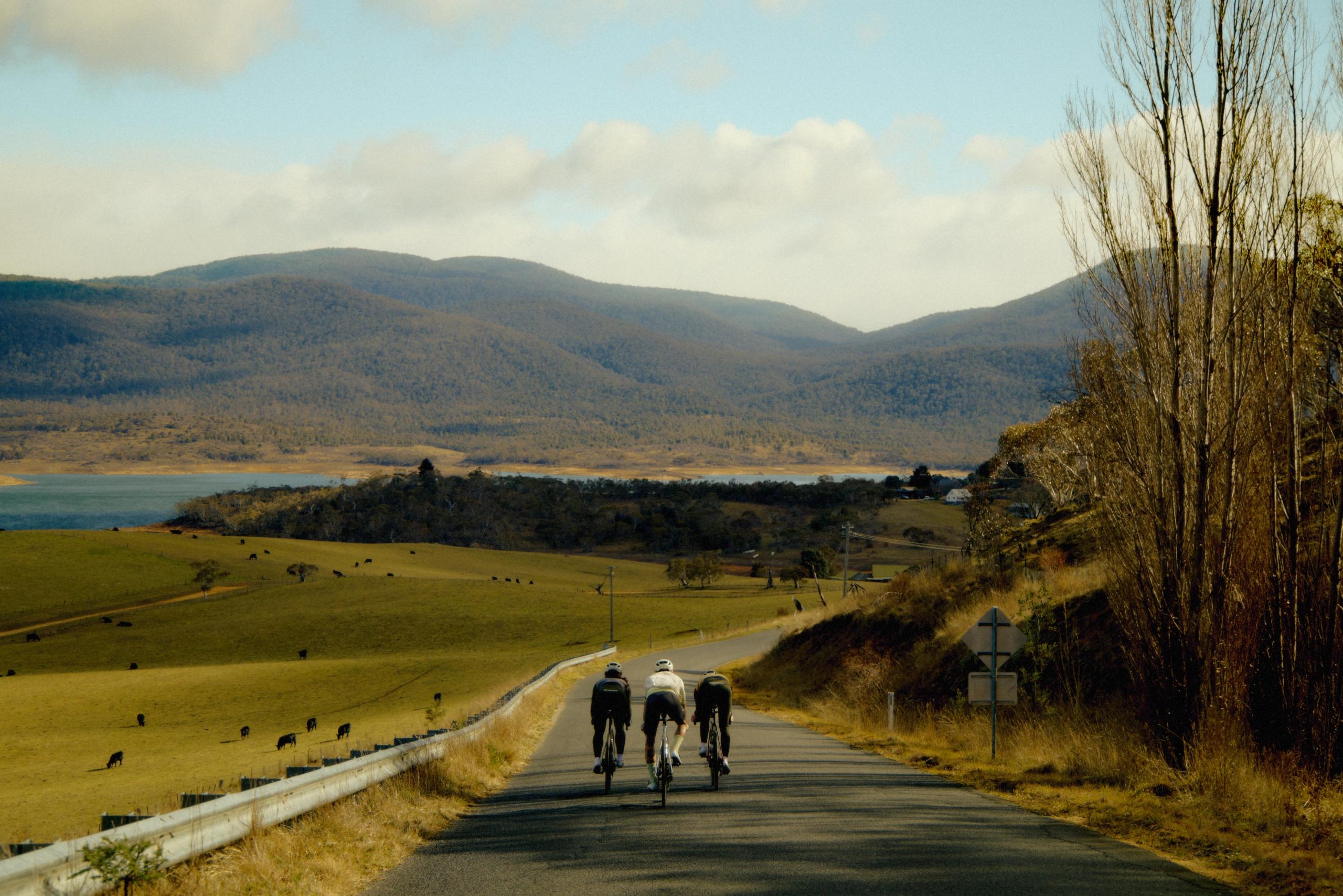 Three cyclists riding on rural road through rolling countryside with mountains and lake in background