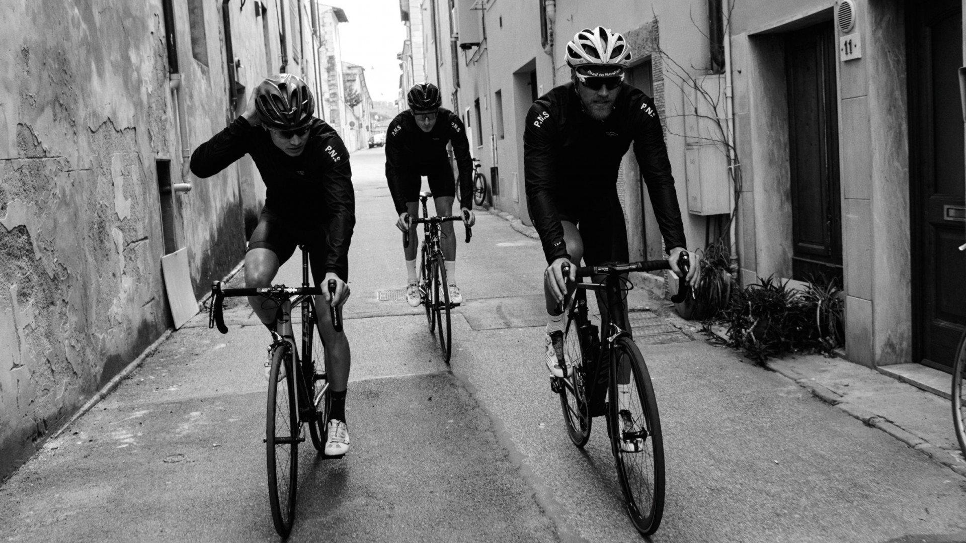 Three cyclists wearing black cycling kit with logo cycling through narrow stone alleyway in black and white