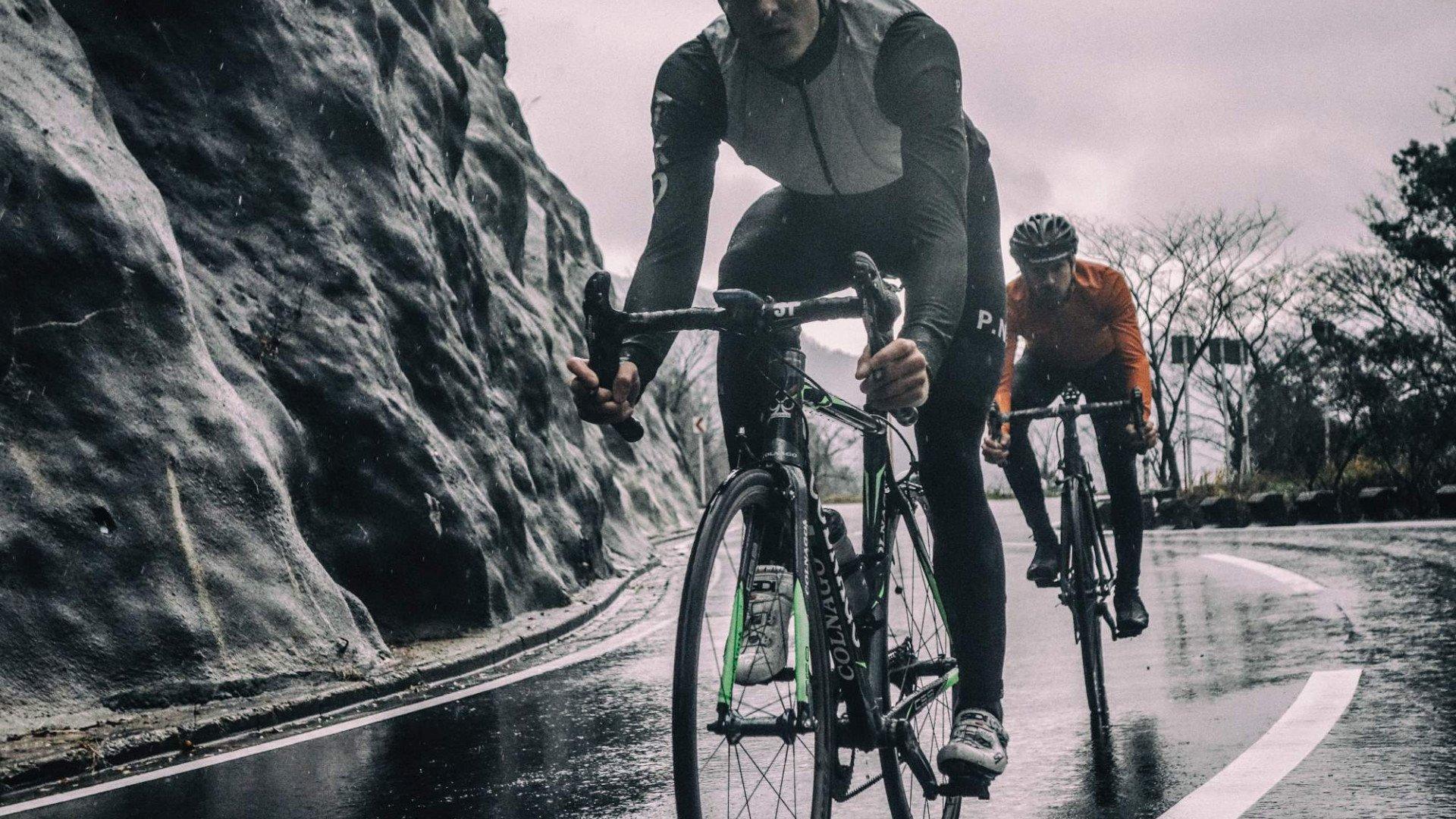 Cyclists riding time trial bikes on wet mountain road alongside rock wall
