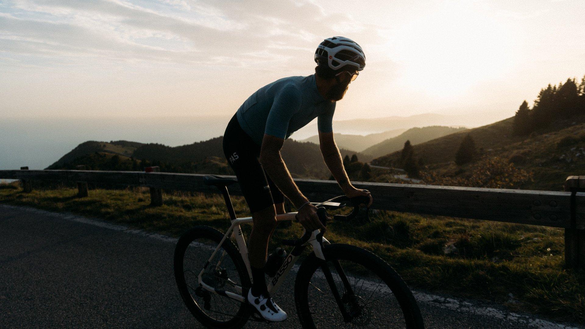 Model wearing dark blue cycling kit in aerodynamic position on time trial bike in mountainous landscape at sunset