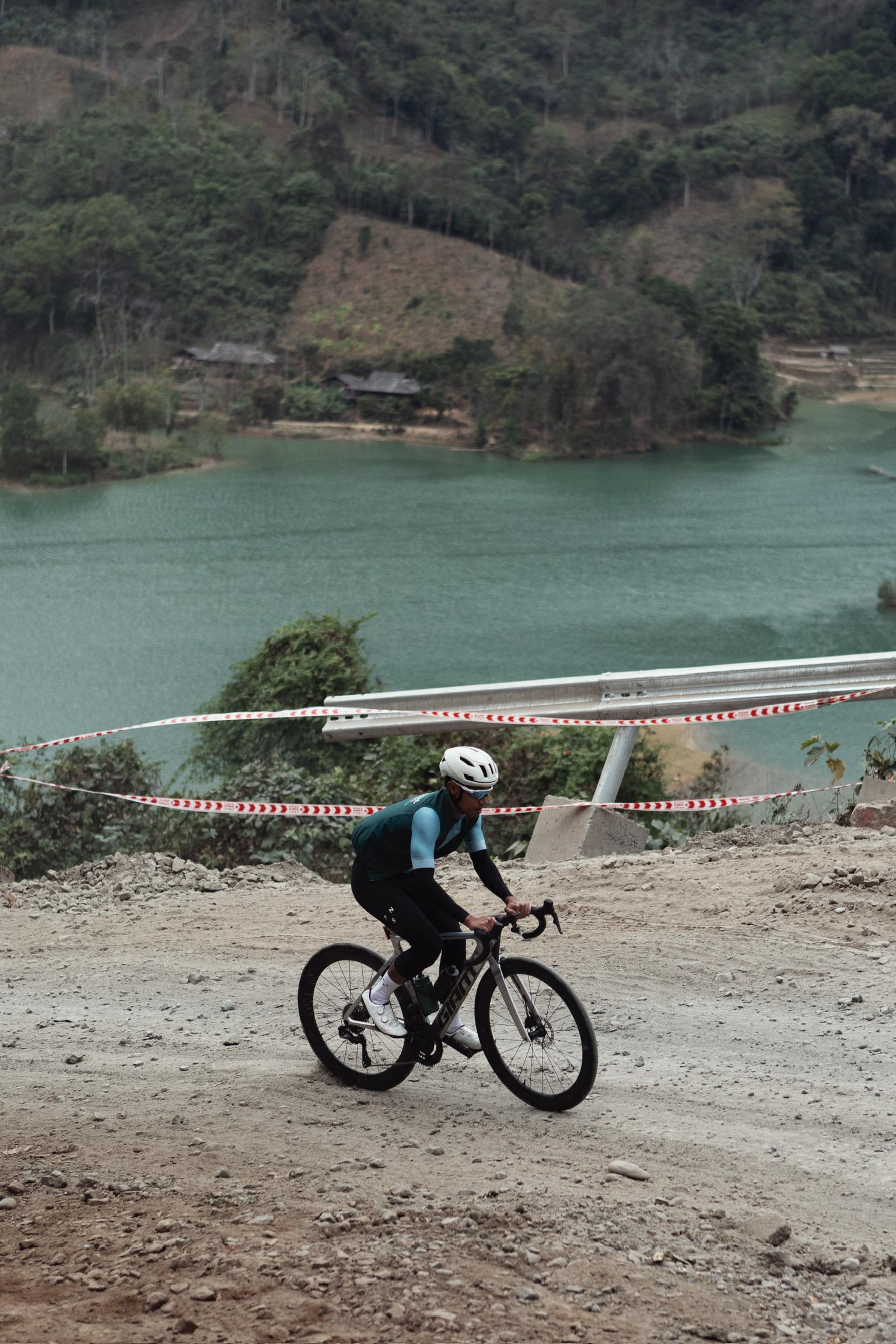 Model wearing green cycling jersey on gravel bike in hilly terrain with lake background