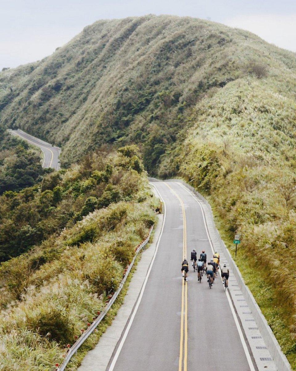 Group of cyclists riding through winding mountain road surrounded by green hills