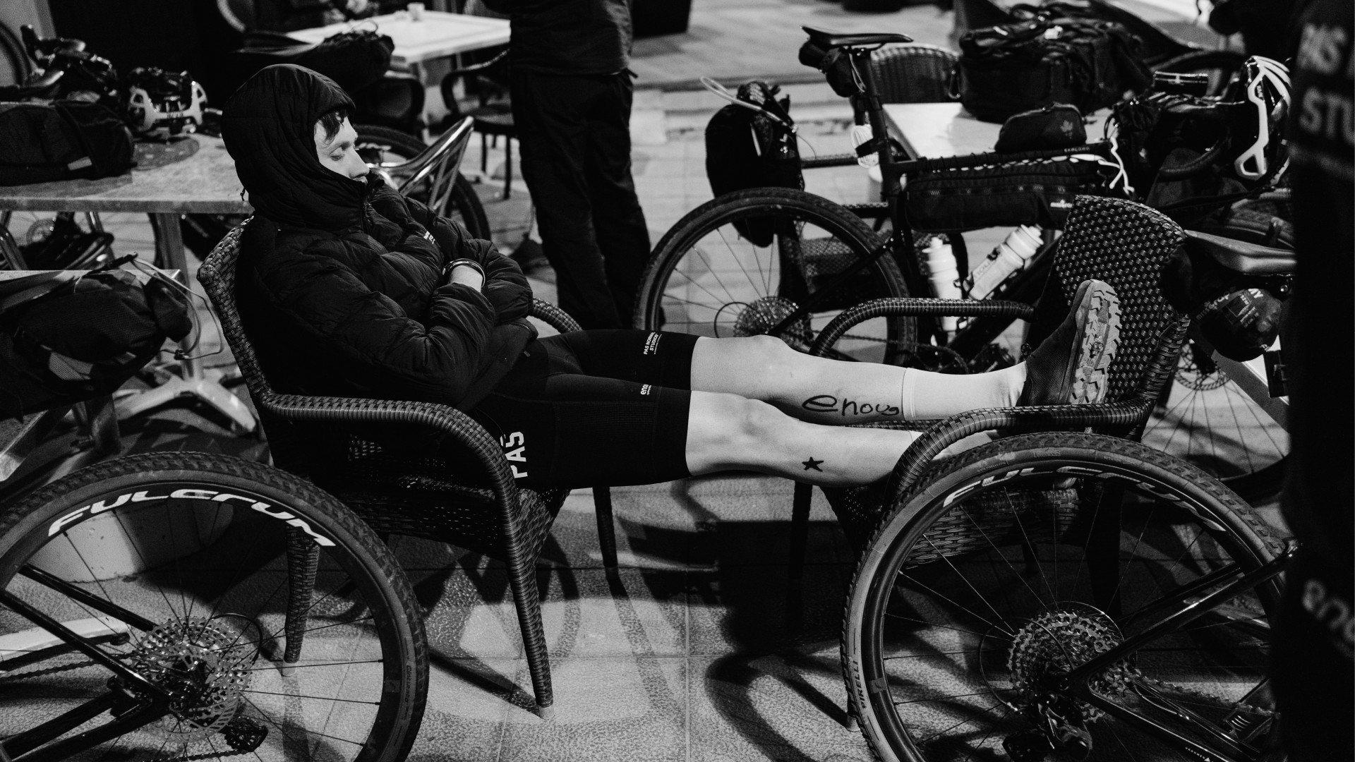 Black and white photo of cyclists resting with bikes at outdoor cafe, model wearing dark cycling apparel