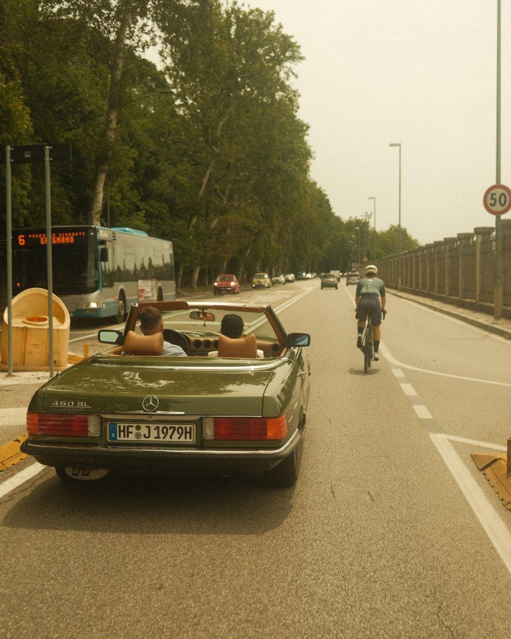Cyclist riding on tree-lined road with vintage Mercedes convertible in foreground
