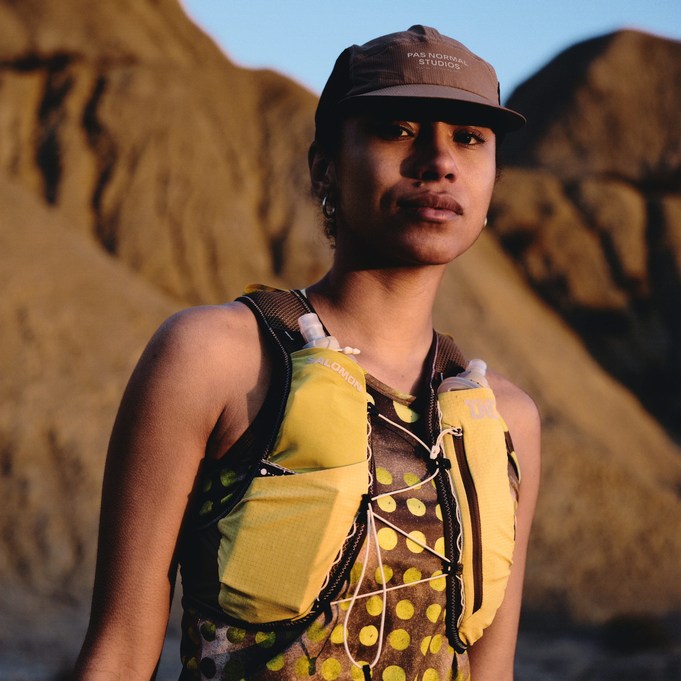 Model wearing yellow patterned running vest with hydration pack and brown cap against desert landscape