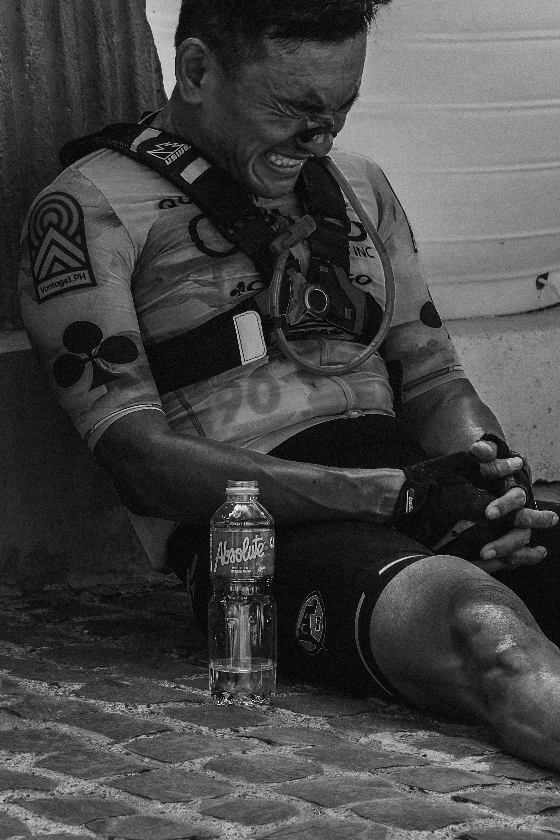 Model wearing cycling jersey with patches and branding, sitting on cobblestones with water bottle, black and white photo