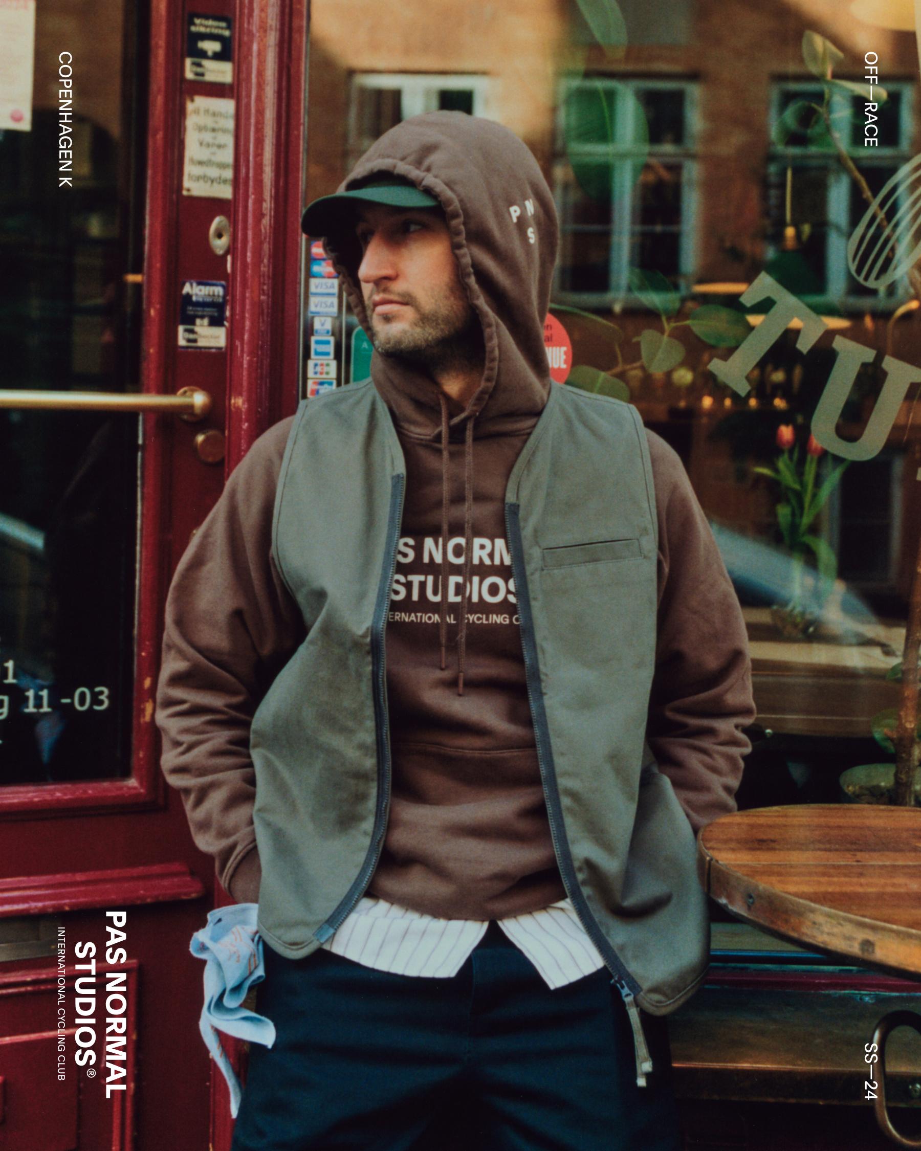 Model wearing Off-Race vest in olive green over brown hoodie with logo, seated outside urban storefront