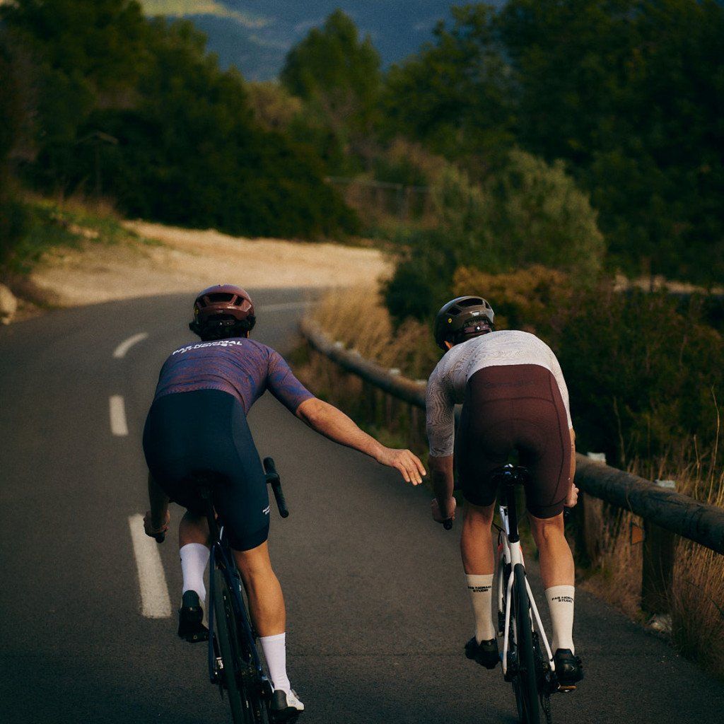 Two cyclists in aero position on road, one wearing purple patterned jersey, other in light colored jersey with dark shorts