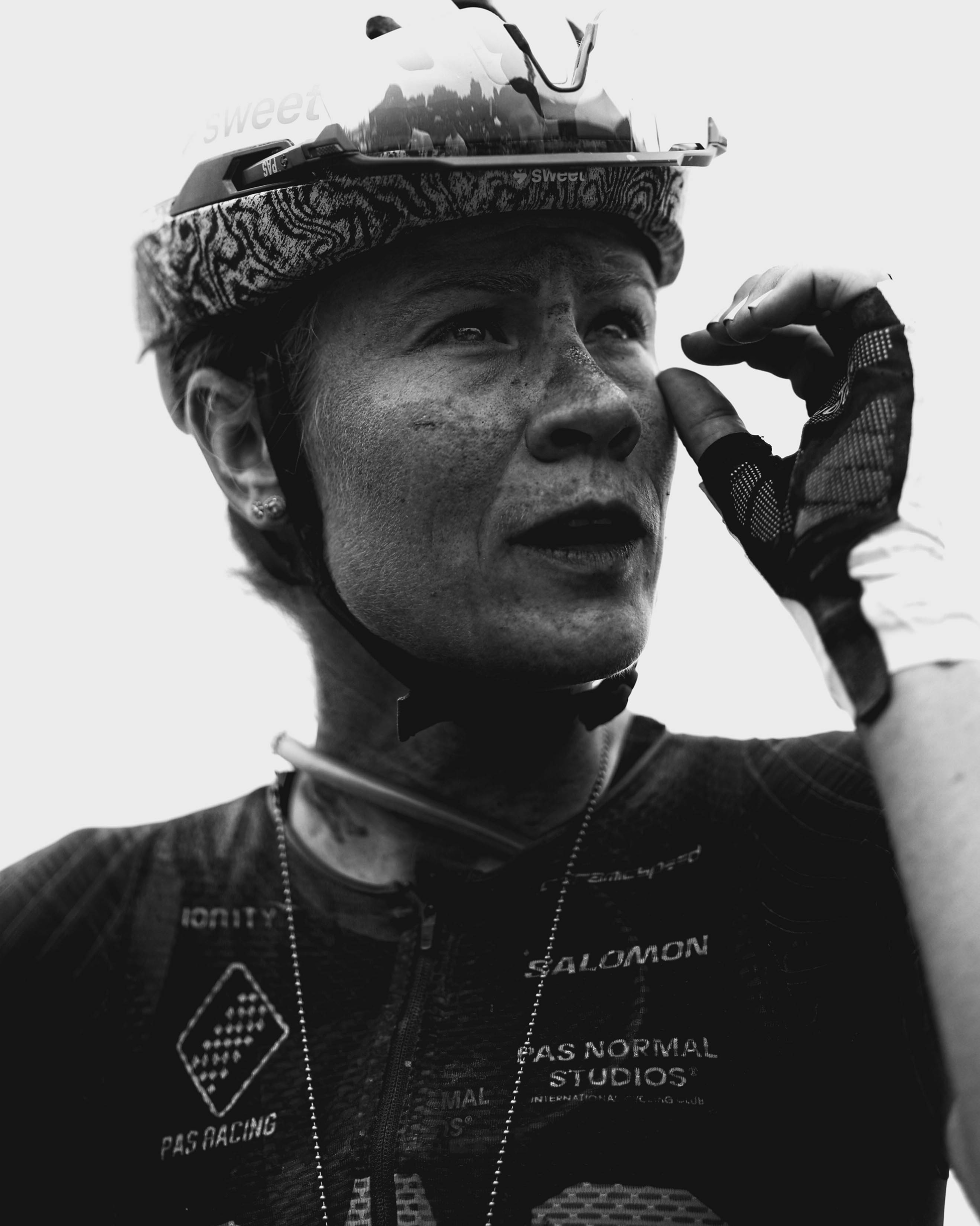 Model wearing black racing jersey with geometric pattern and cycling cap, adjusting helmet strap in black and white portrait