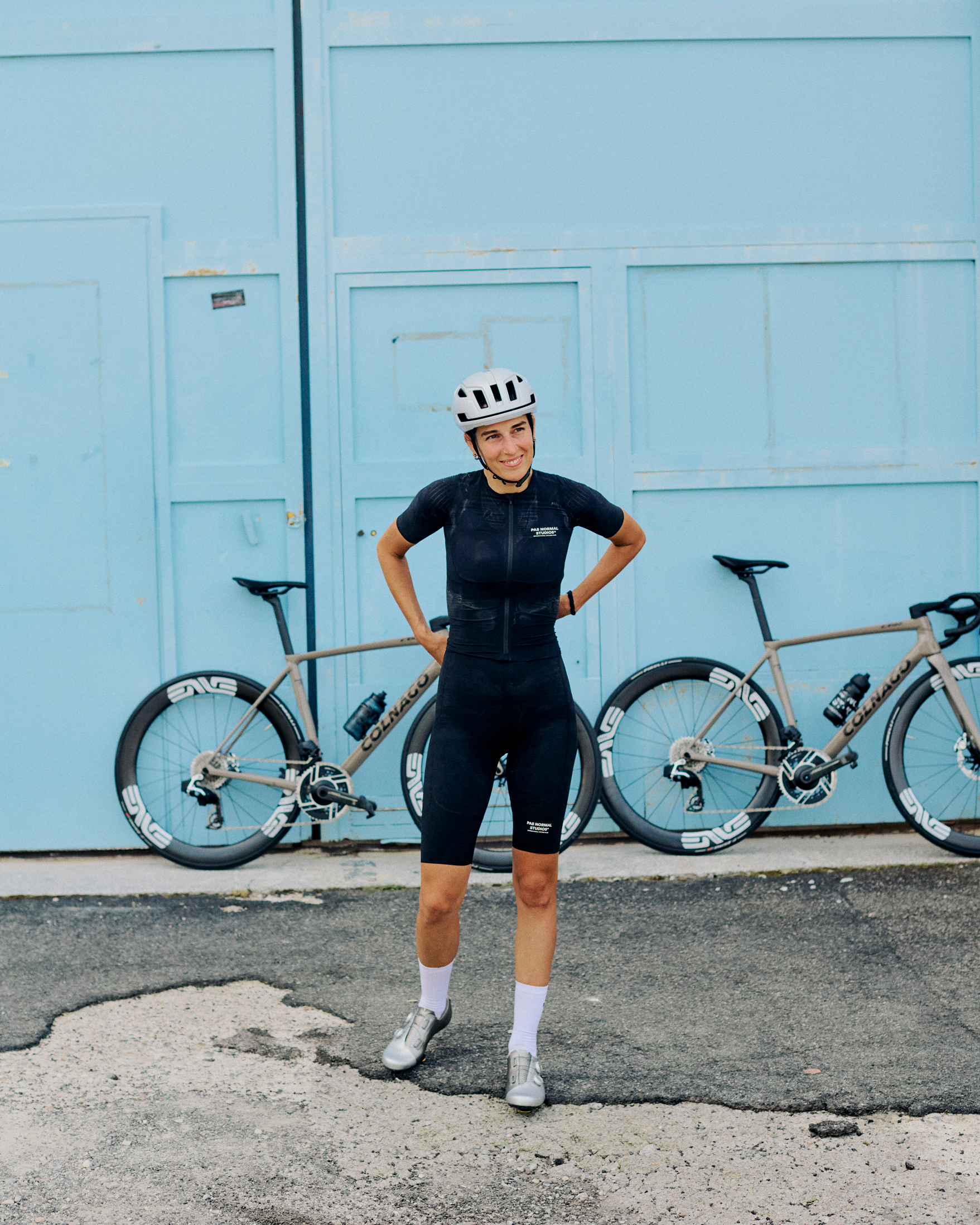 Model wearing black cycling skinsuit with white helmet standing next to Colnago road bike against blue doors