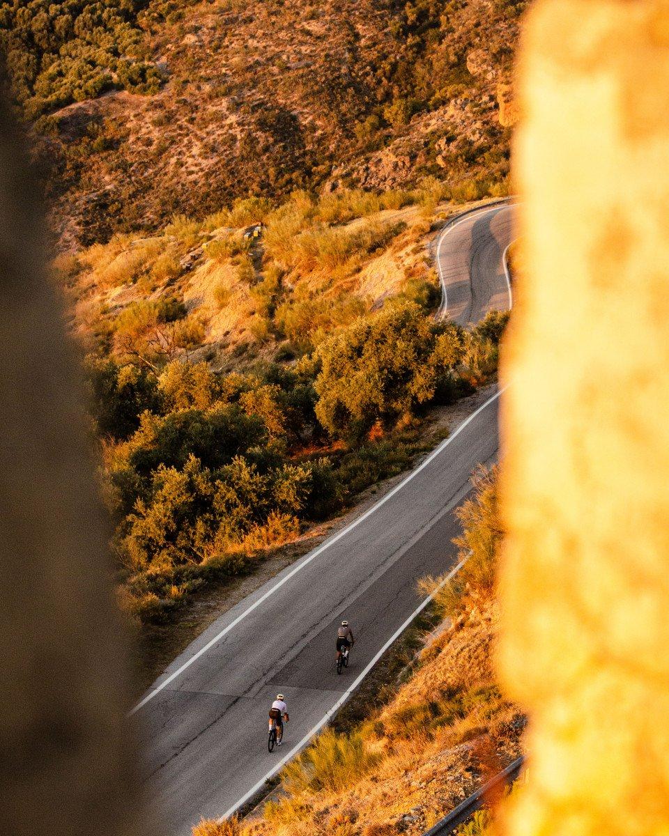Two cyclists on winding mountain road through golden autumn landscape at sunset