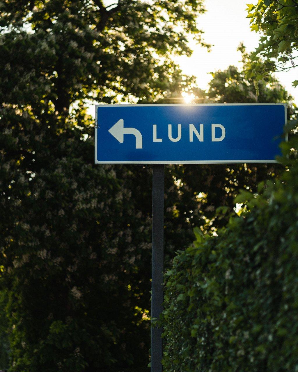 Blue road sign pointing to Lund surrounded by green trees with sunlight filtering through