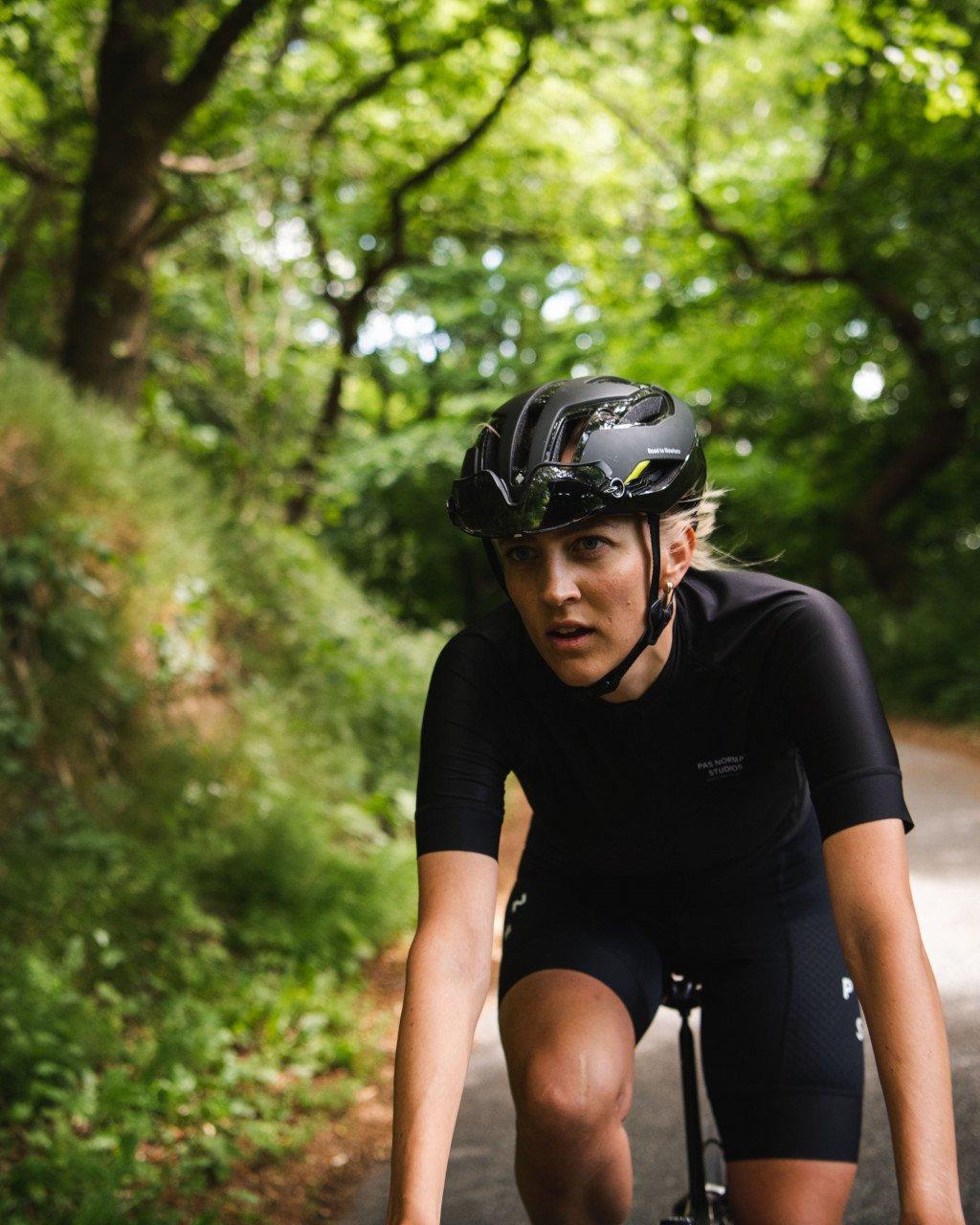 Model wearing black cycling jersey and shorts with aerodynamic helmet on tree-lined road
