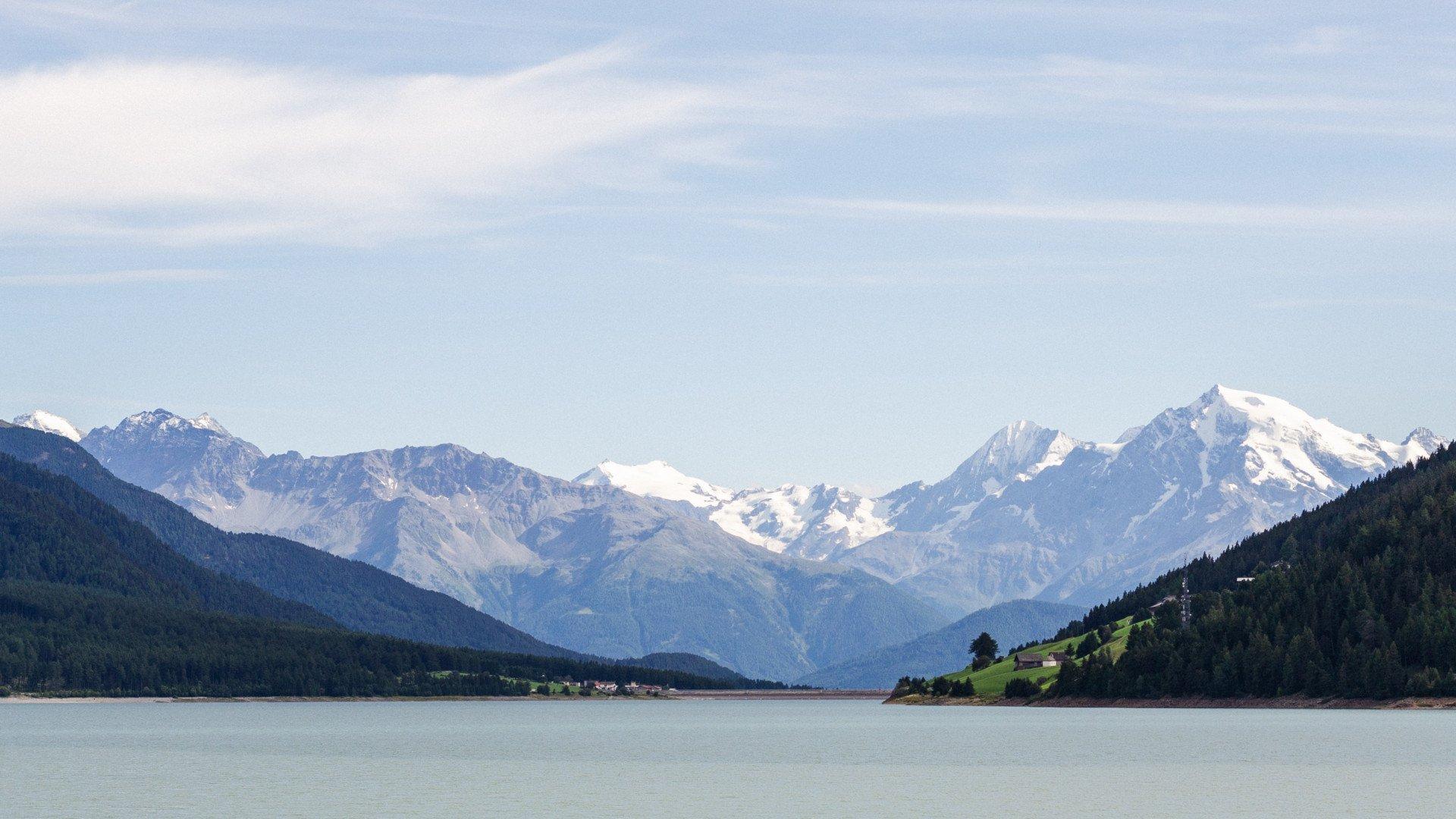Alpine landscape with snow-capped mountains, forested hills and lake under cloudy sky