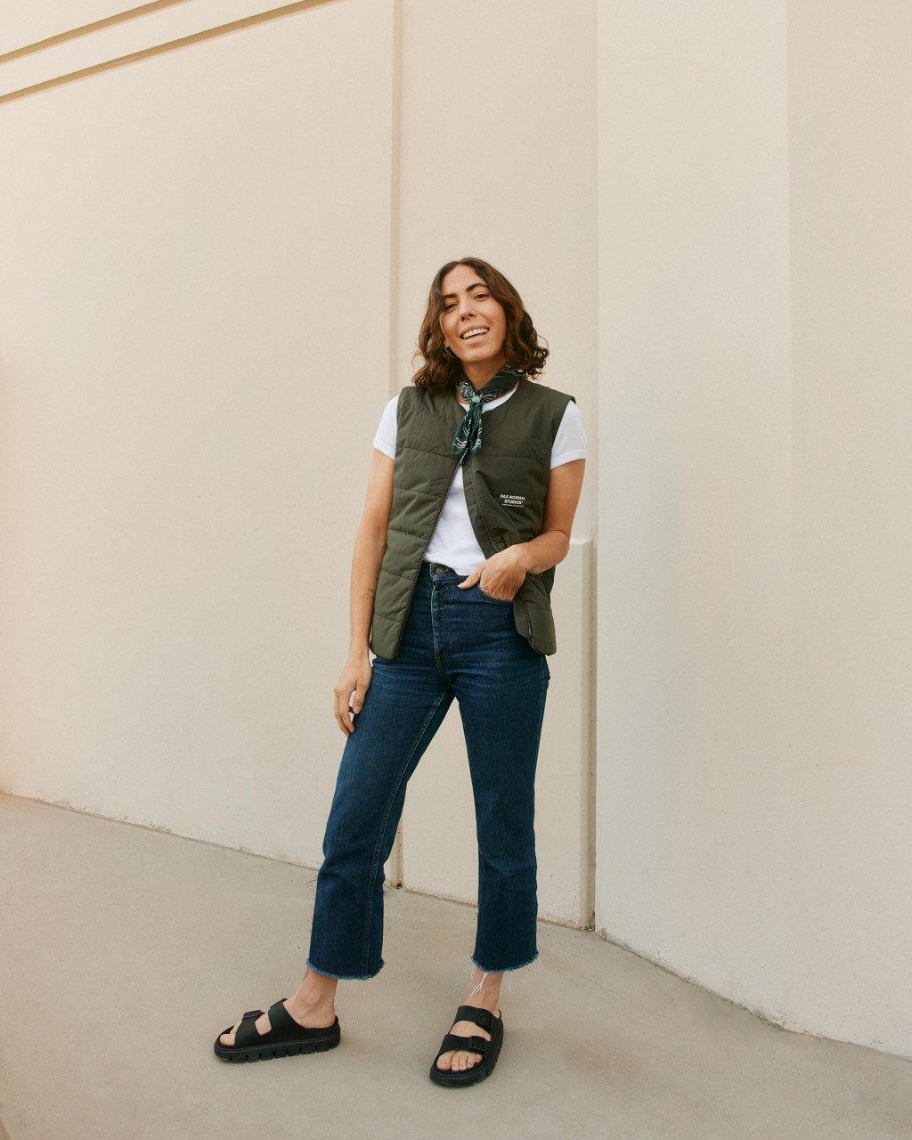Model wearing olive green vest with white t-shirt and jeans, smiling against beige wall