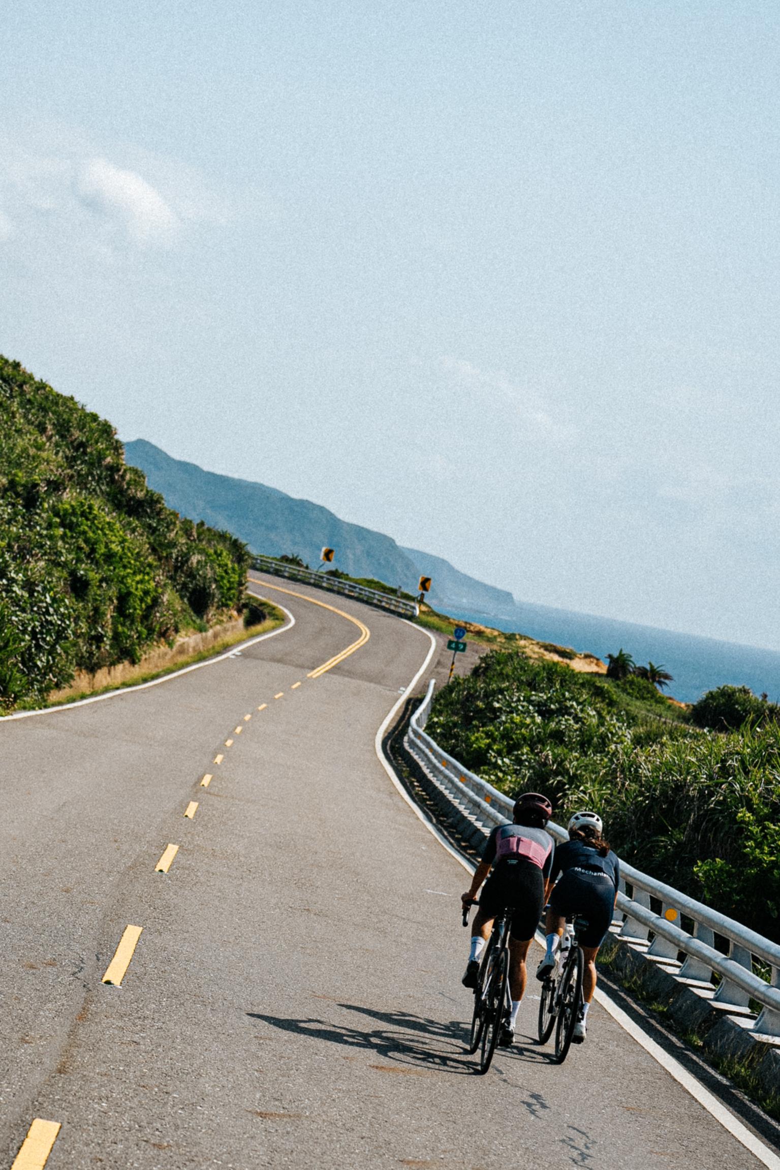 Two cyclists riding on winding coastal road with ocean and mountains in background