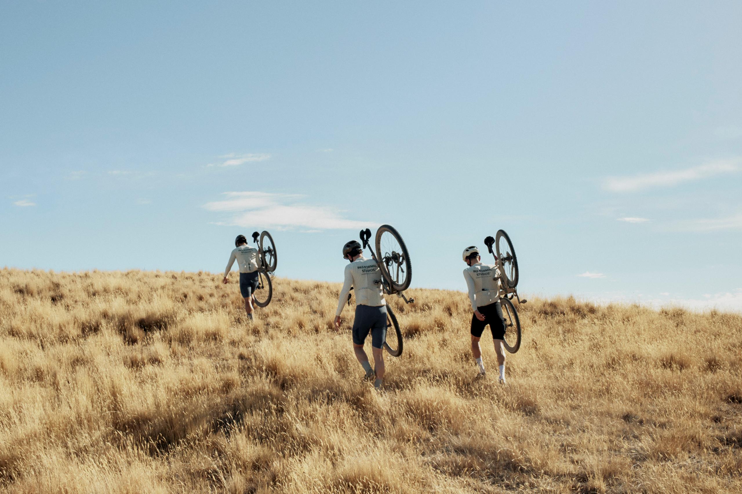 Three cyclists carrying bikes across golden grassland under blue sky