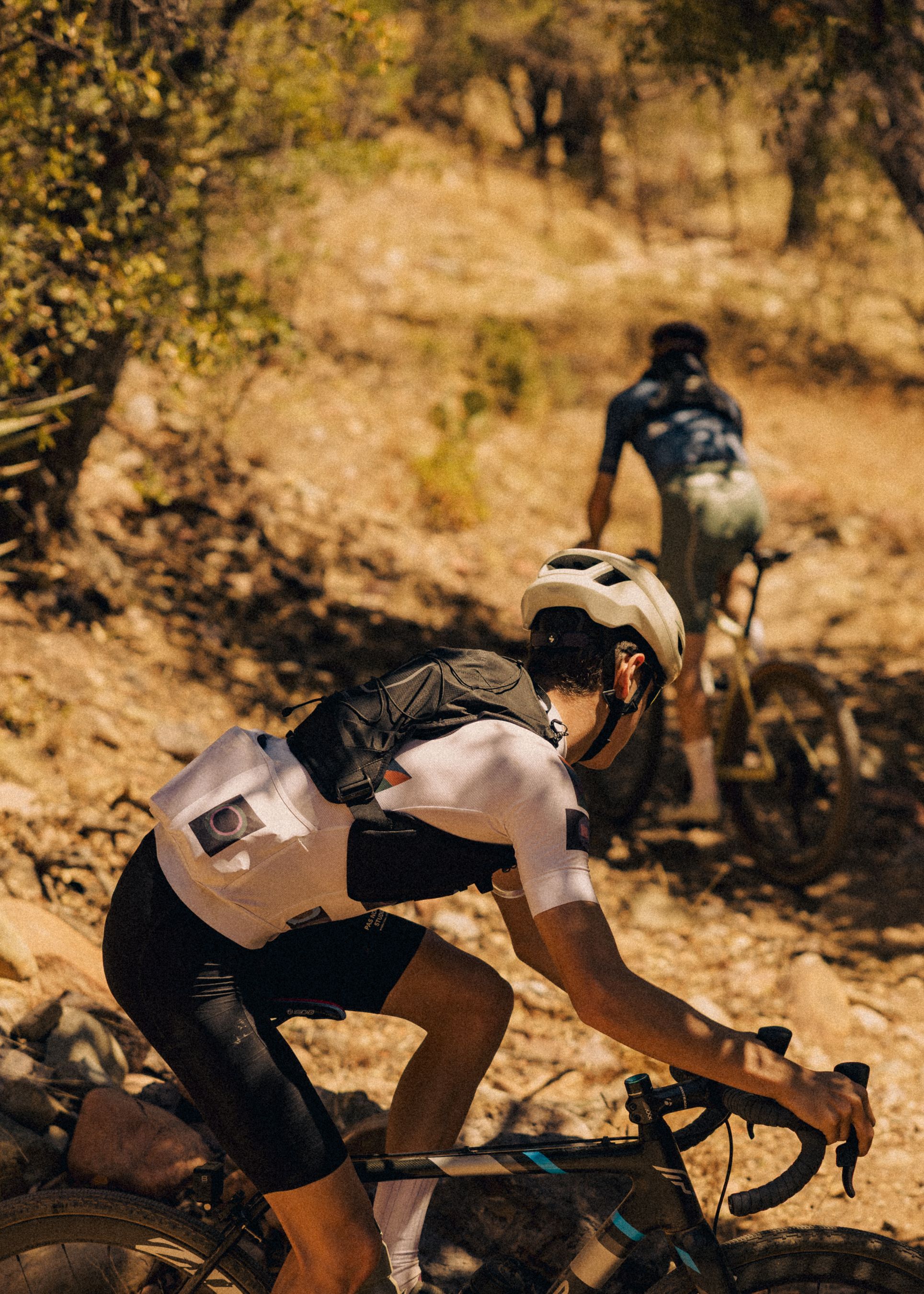 Model wearing light grey cycling jersey and black shorts mountain biking on dirt trail through wooded terrain