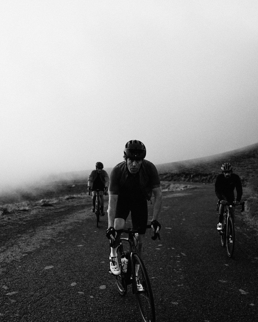 Three cyclists riding on desert road in black and white, misty mountainous landscape in background
