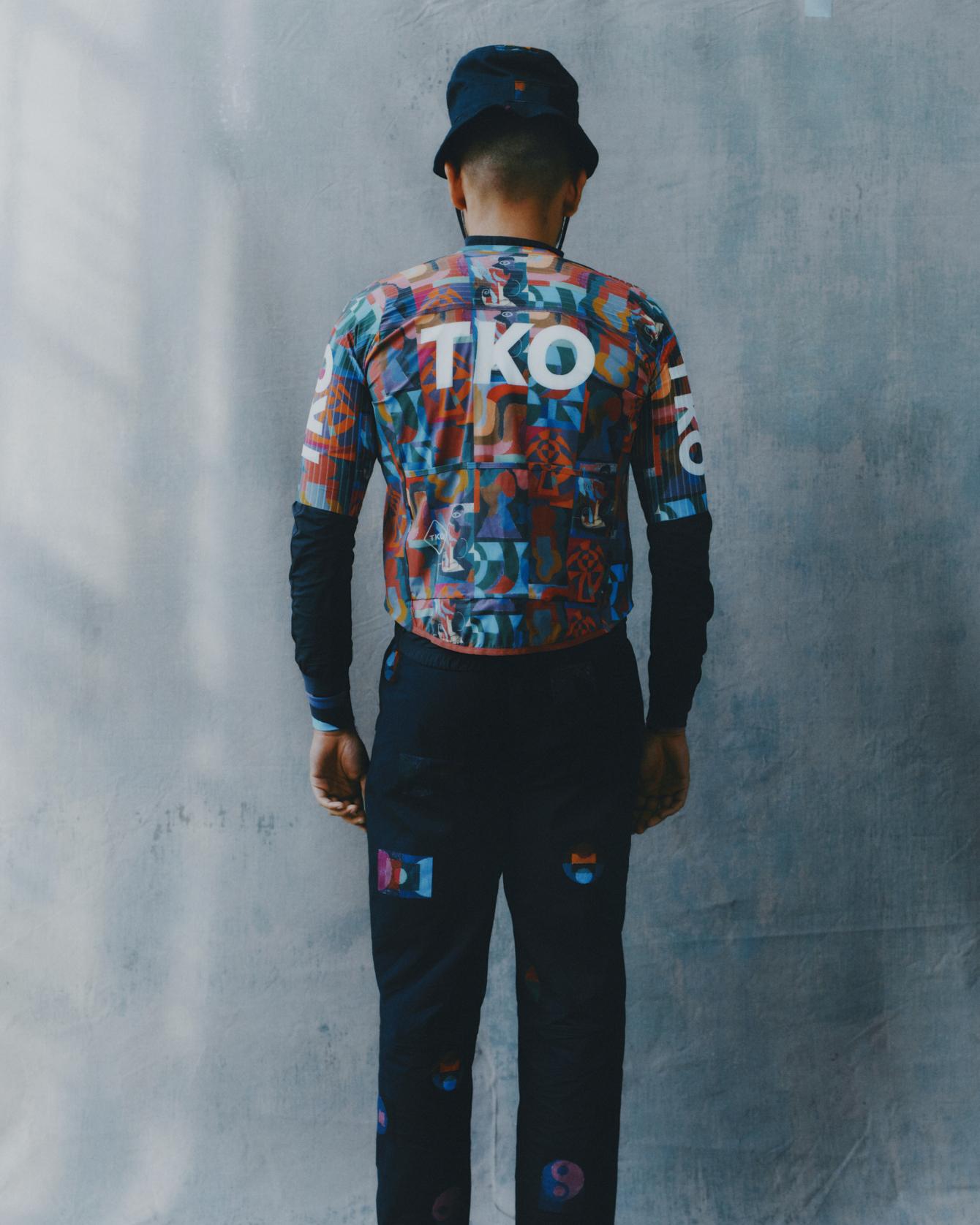 Model wearing T.K.O. jersey in multicolored collage print with black arm warmers and black trousers, back view