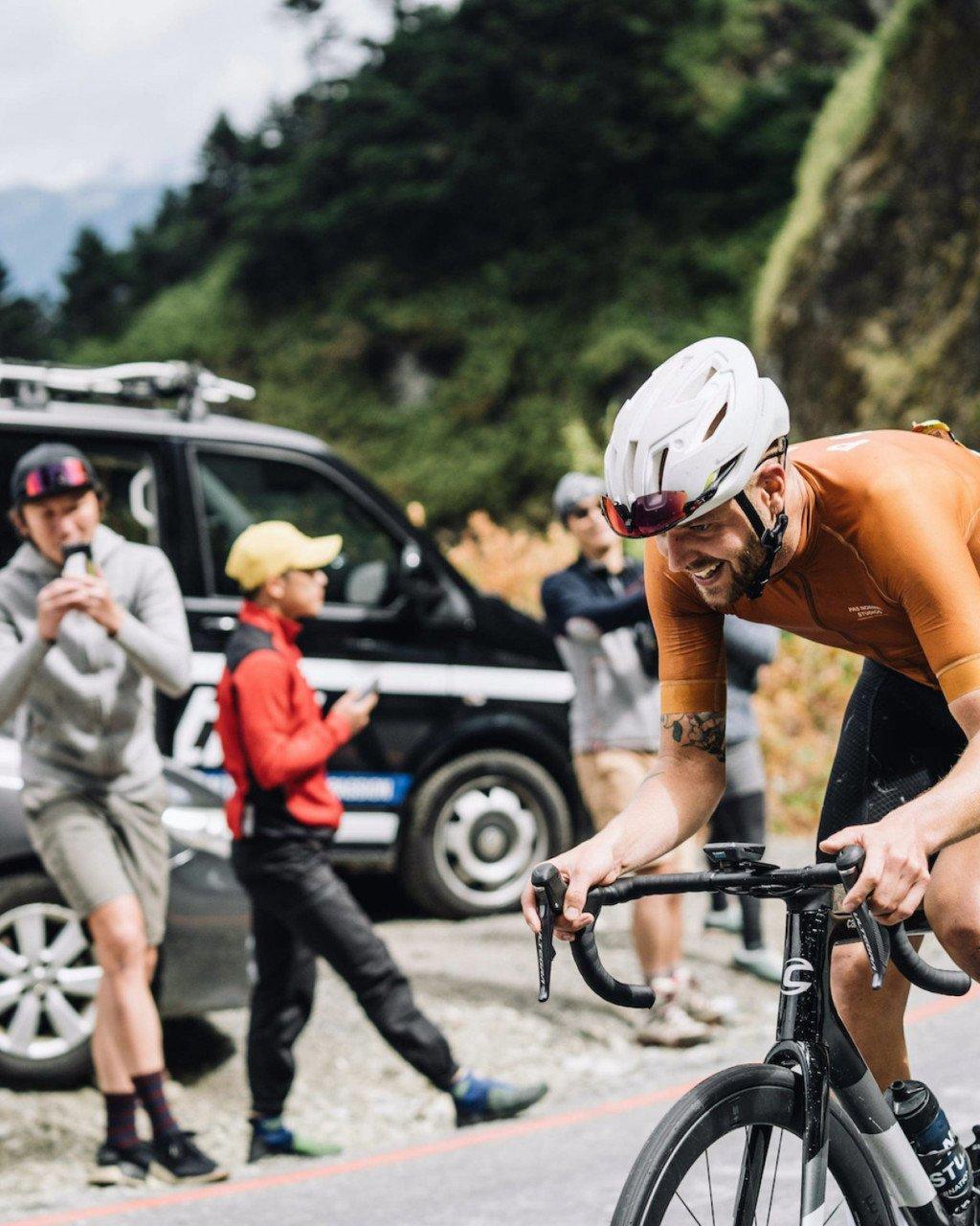 Model wearing orange time trial jersey and white helmet during race with spectators and support vehicles