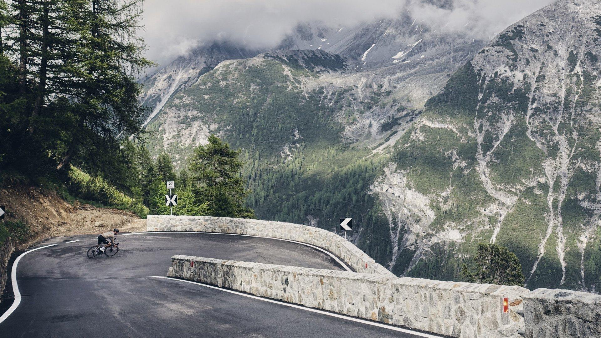 Cyclist on winding mountain road with dramatic Alpine peaks and switchback turns