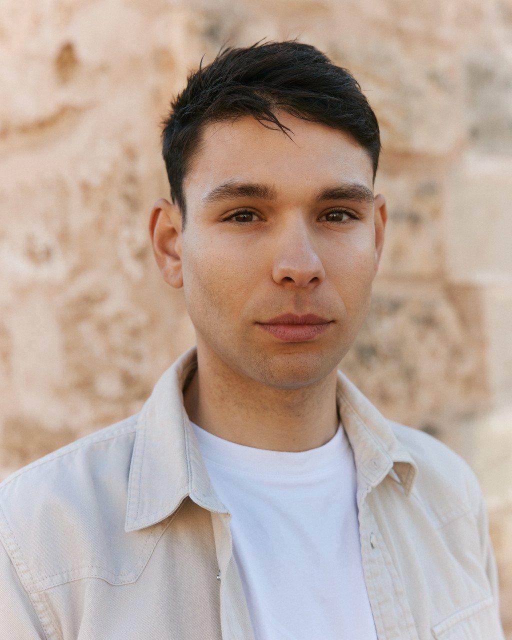 Model wearing white t-shirt and light button-up shirt against stone wall background