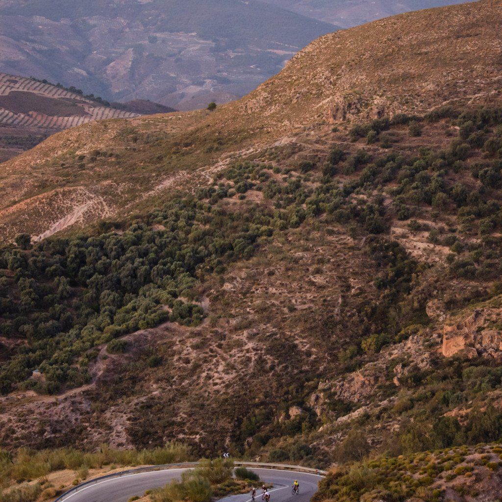 Two cyclists on winding mountain road through golden hour landscape with rolling hills and Mediterranean terrain