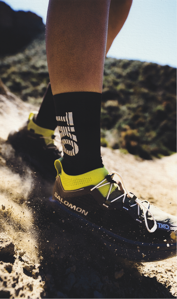 Model wearing black cycling socks with logo branding and yellow Salomon running shoes on trail