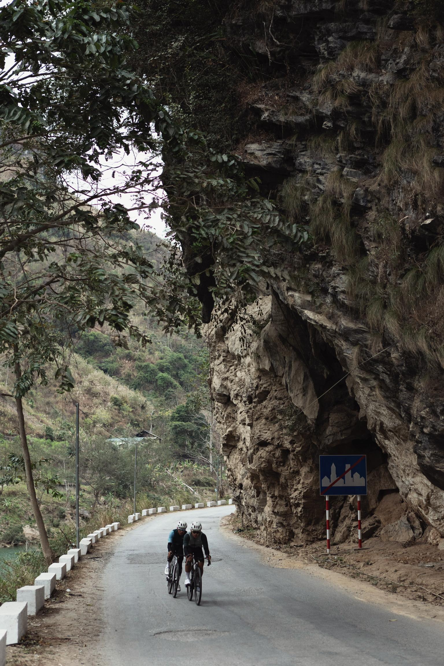 Two cyclists riding on mountain road through rocky terrain with moss-covered cliffs