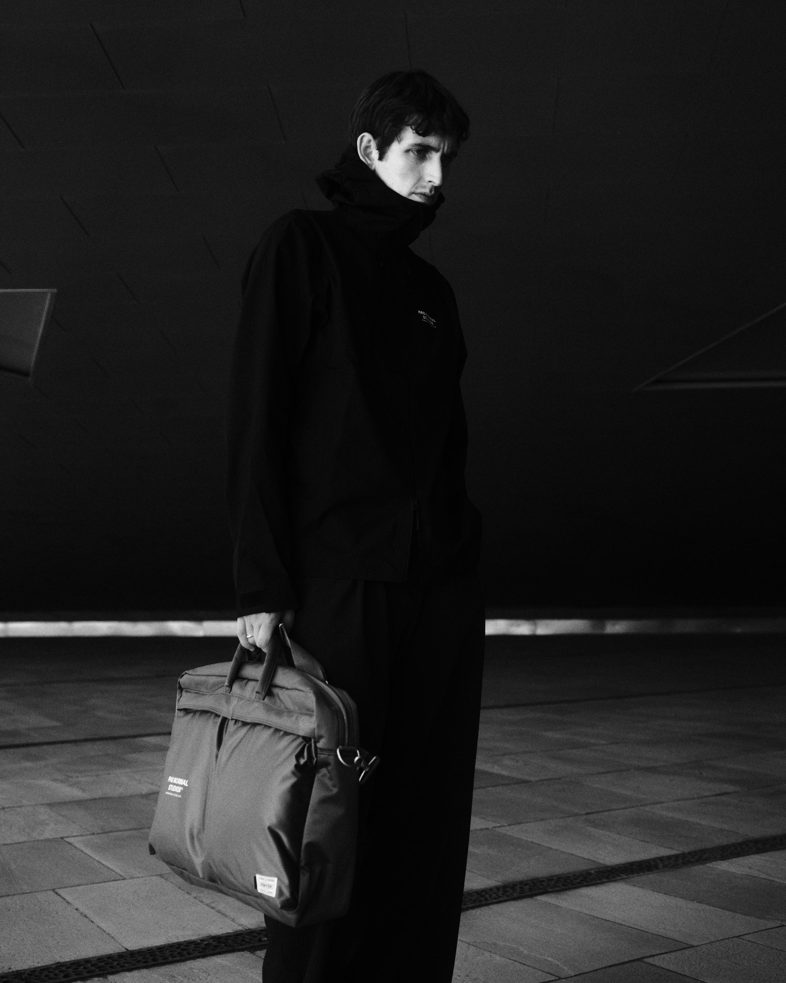 Model wearing black hooded jacket holding grey Porter-Yoshida collaboration bag in urban setting