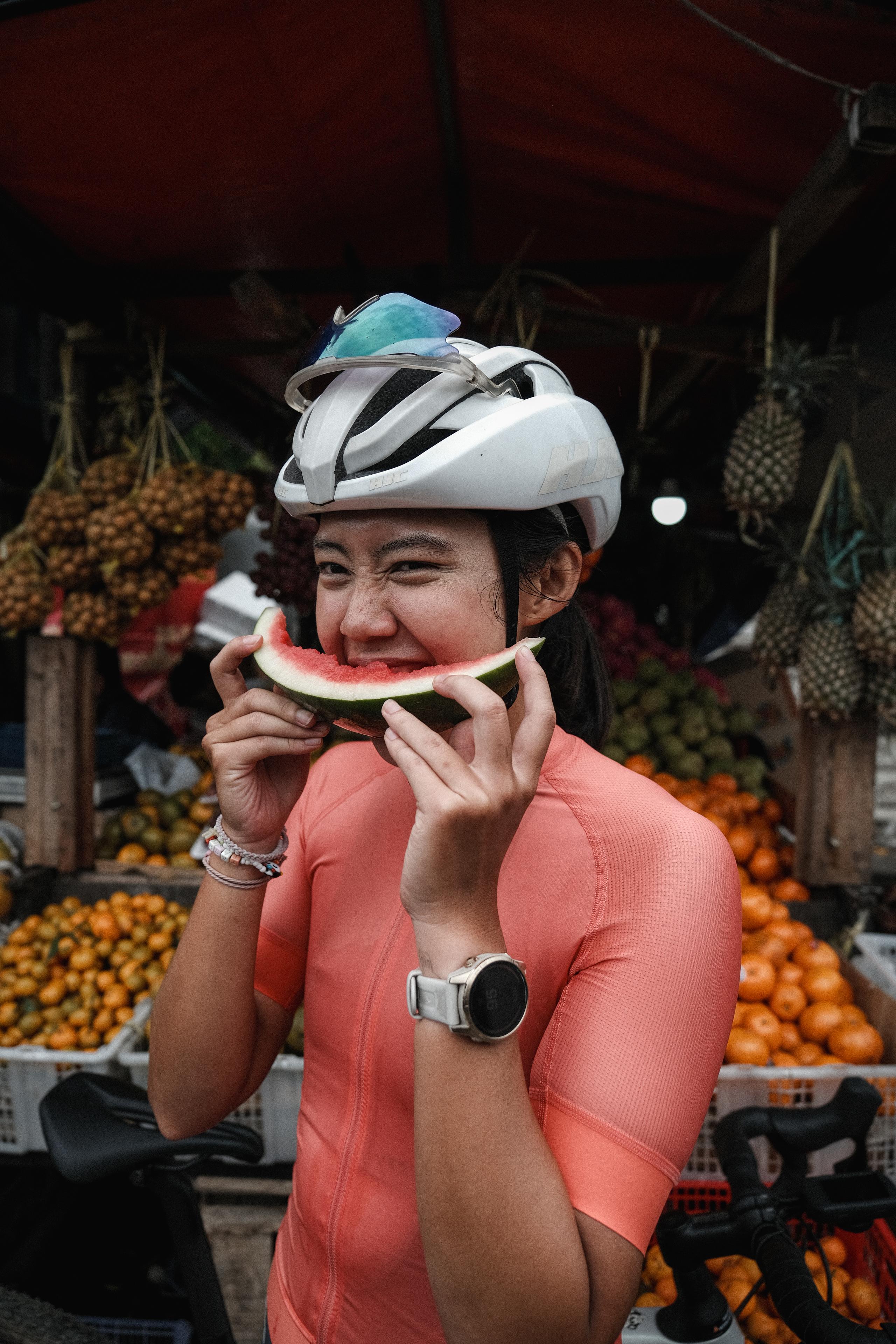 Model wearing coral long-sleeve cycling top eating watermelon at fruit market
