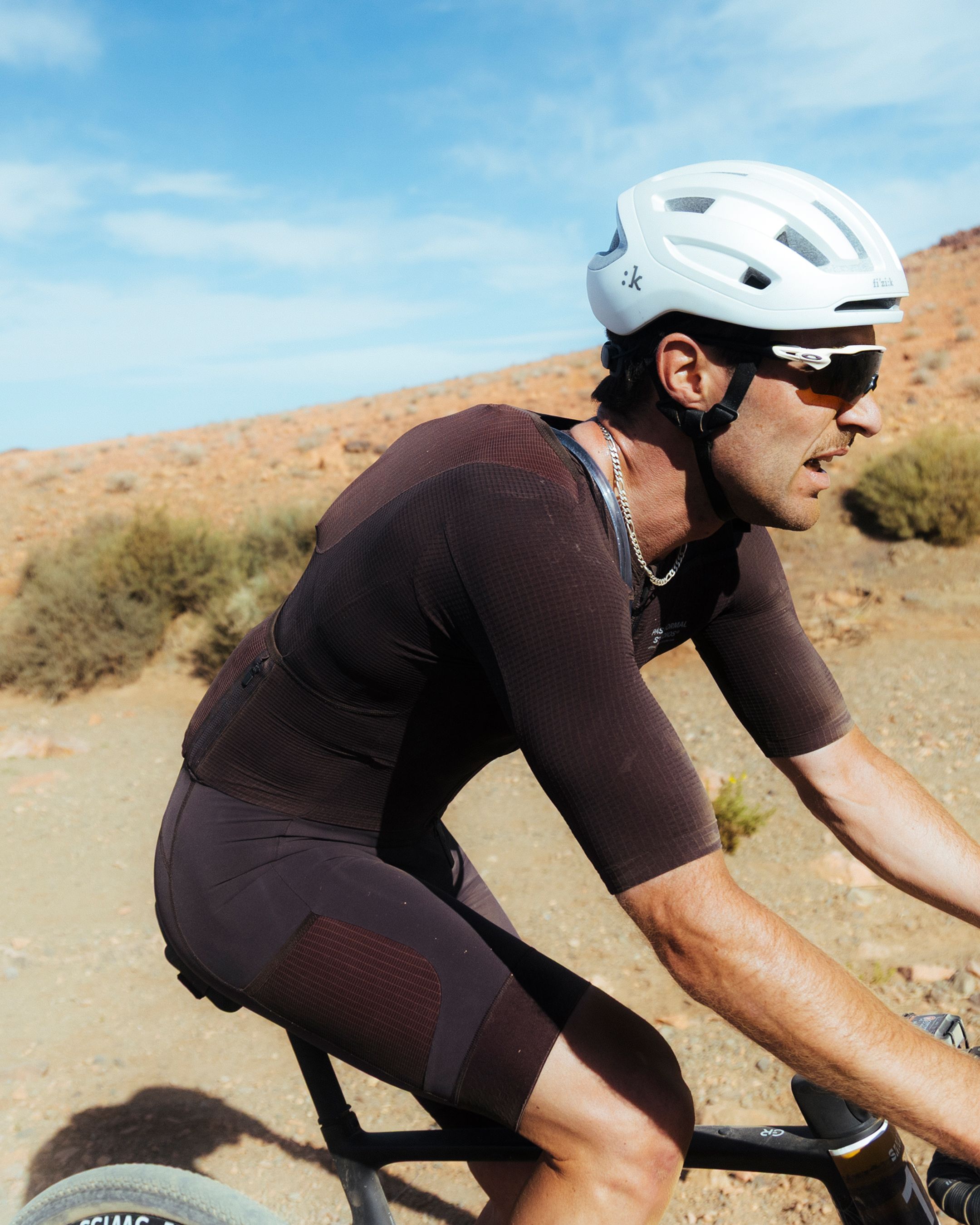 Model wearing Men's Mechanism Jersey in brown cycling in desert terrain with white helmet and sunglasses