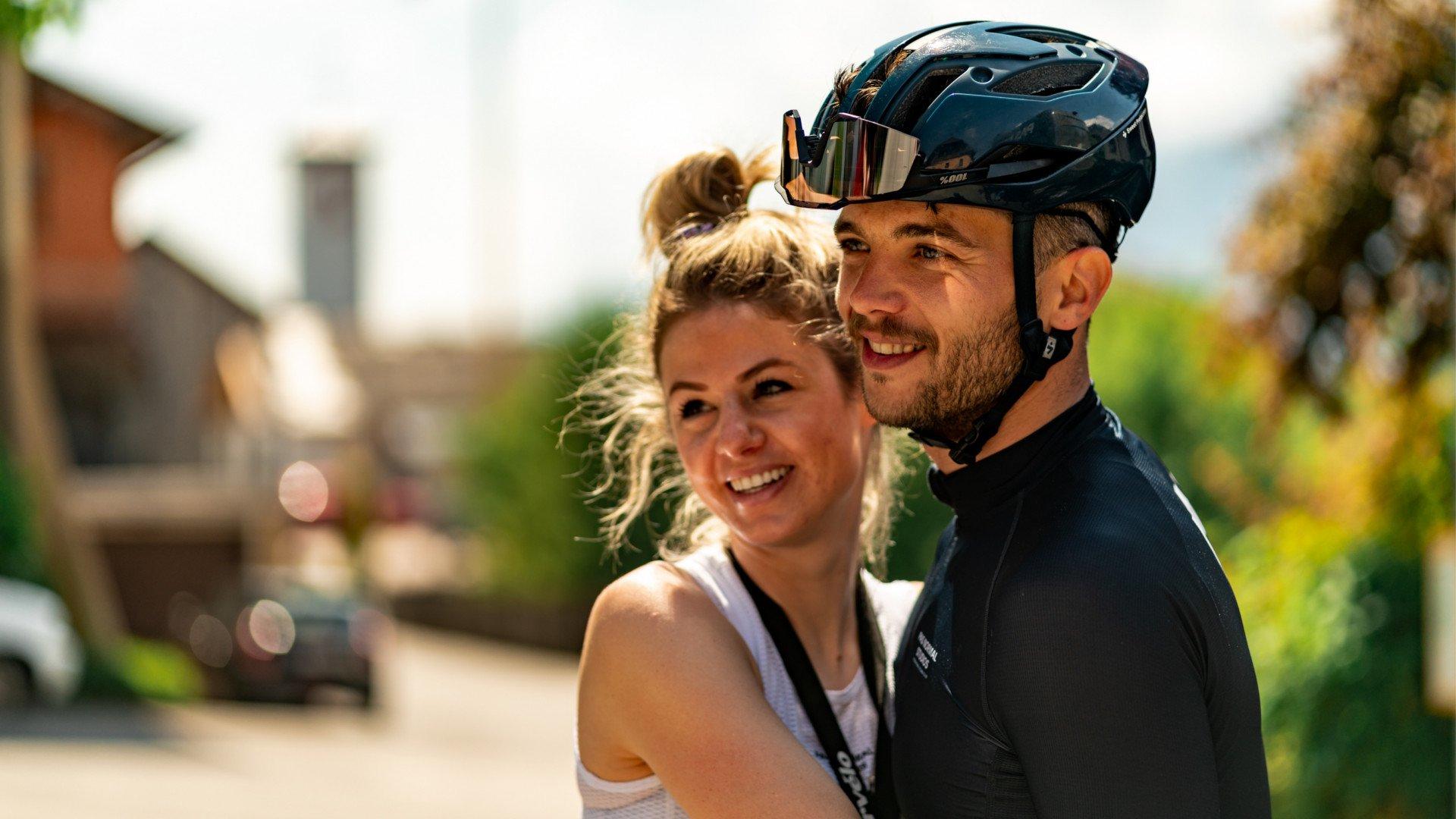 Model wearing black cycling jersey with helmet and sunglasses embracing woman in white tank top outdoors