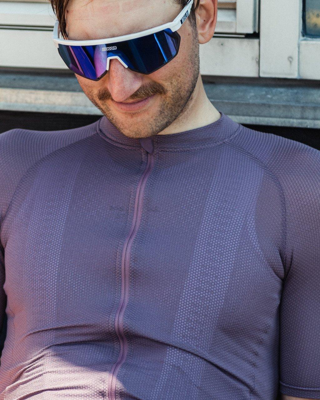 Model wearing purple cycling jersey with textured fabric and partial zip, blue mirrored sunglasses