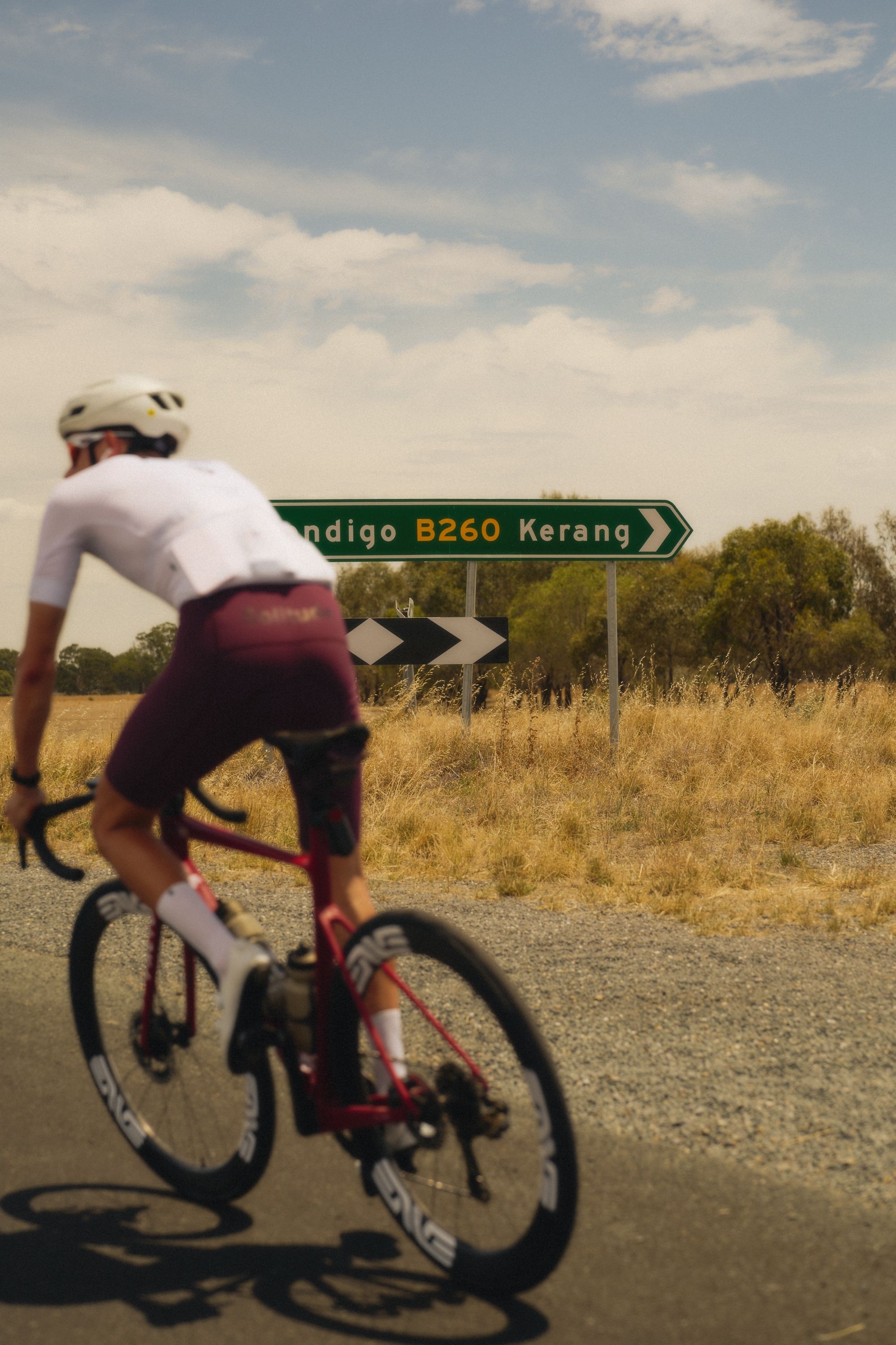 Model wearing white cycling jersey and red shorts cycling on rural road past road sign