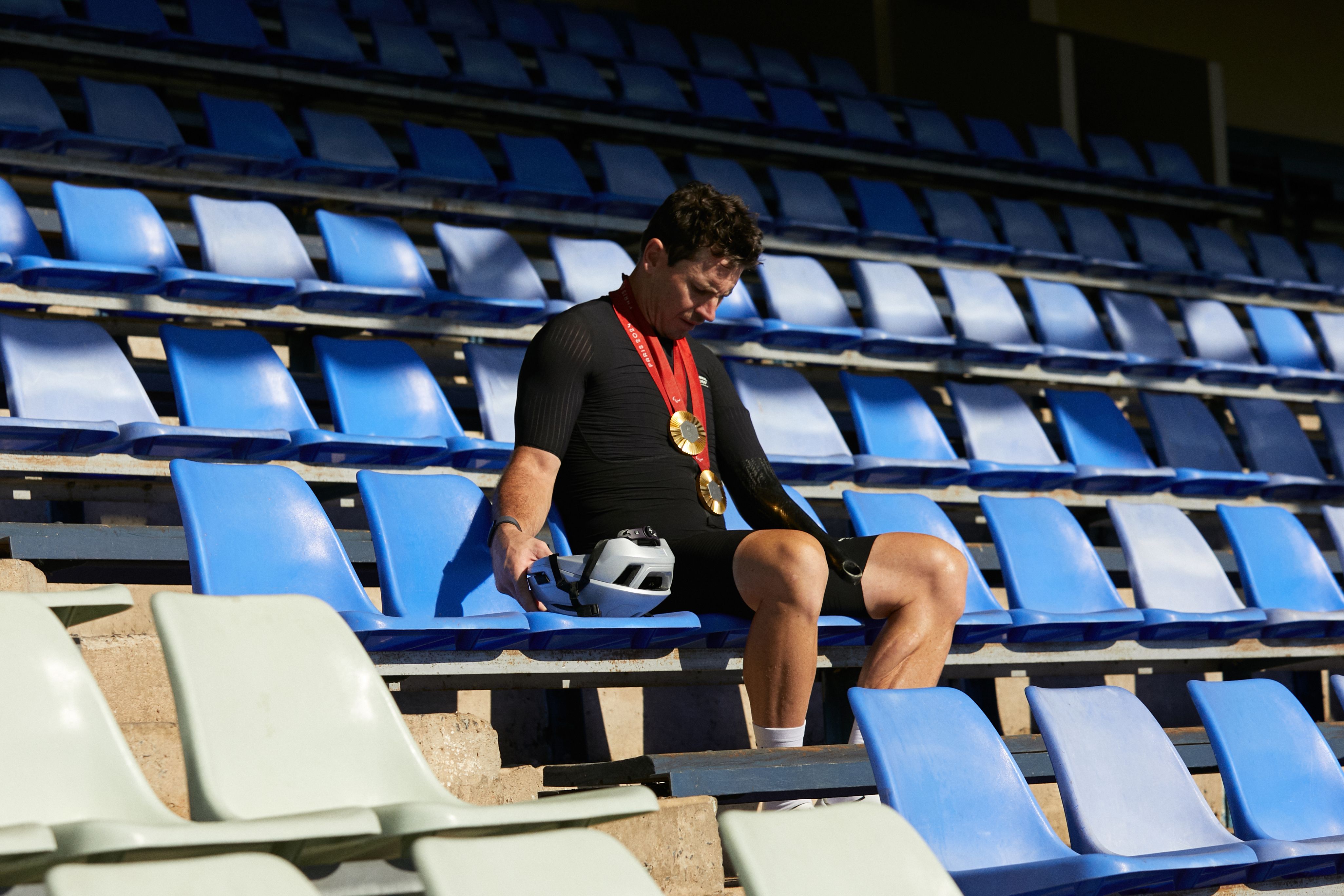 Model wearing black cycling jersey and shorts in stadium seating, holding helmet with medals around neck