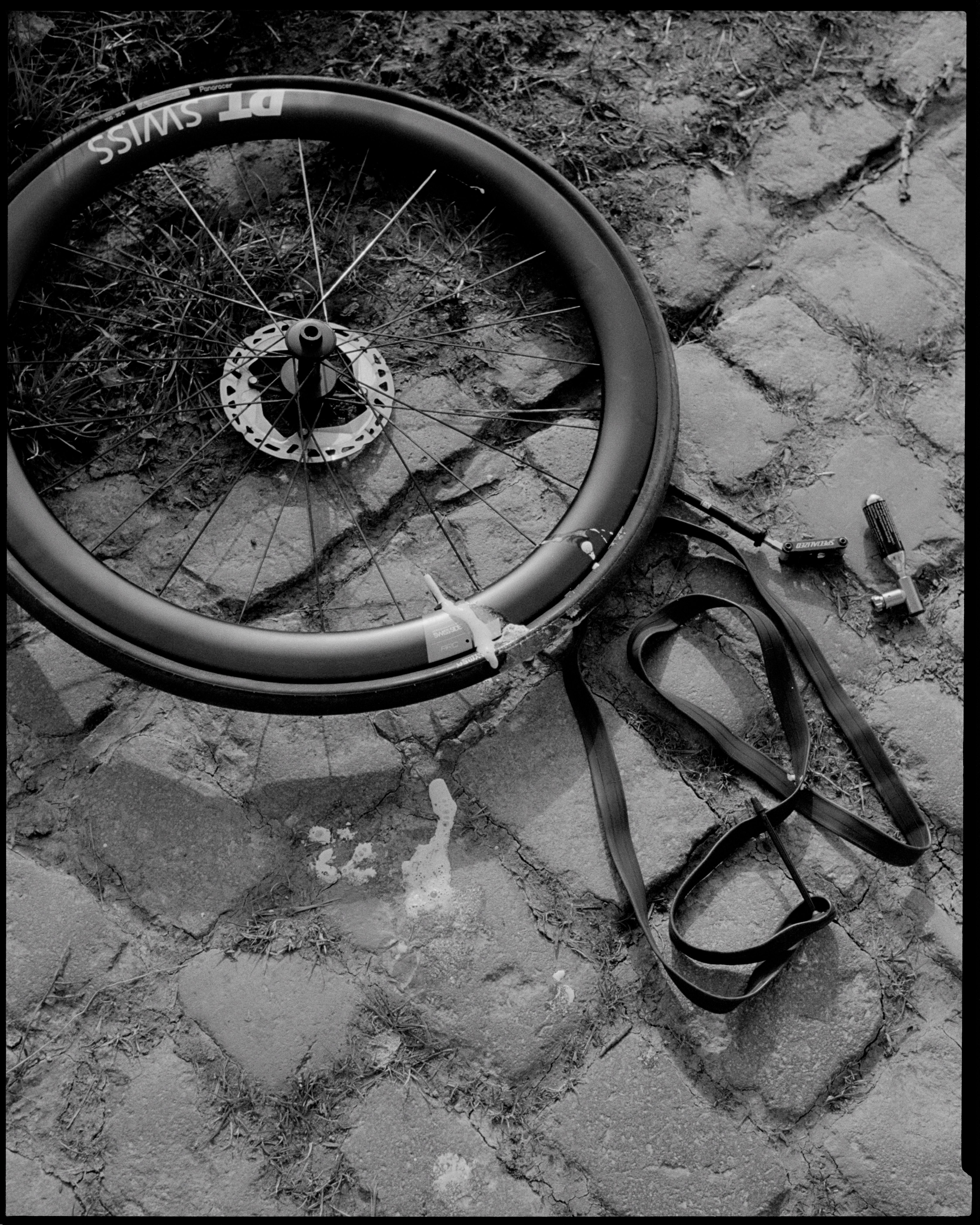 Black and white photo of bicycle wheel with disc brake and pedals on cobblestone street