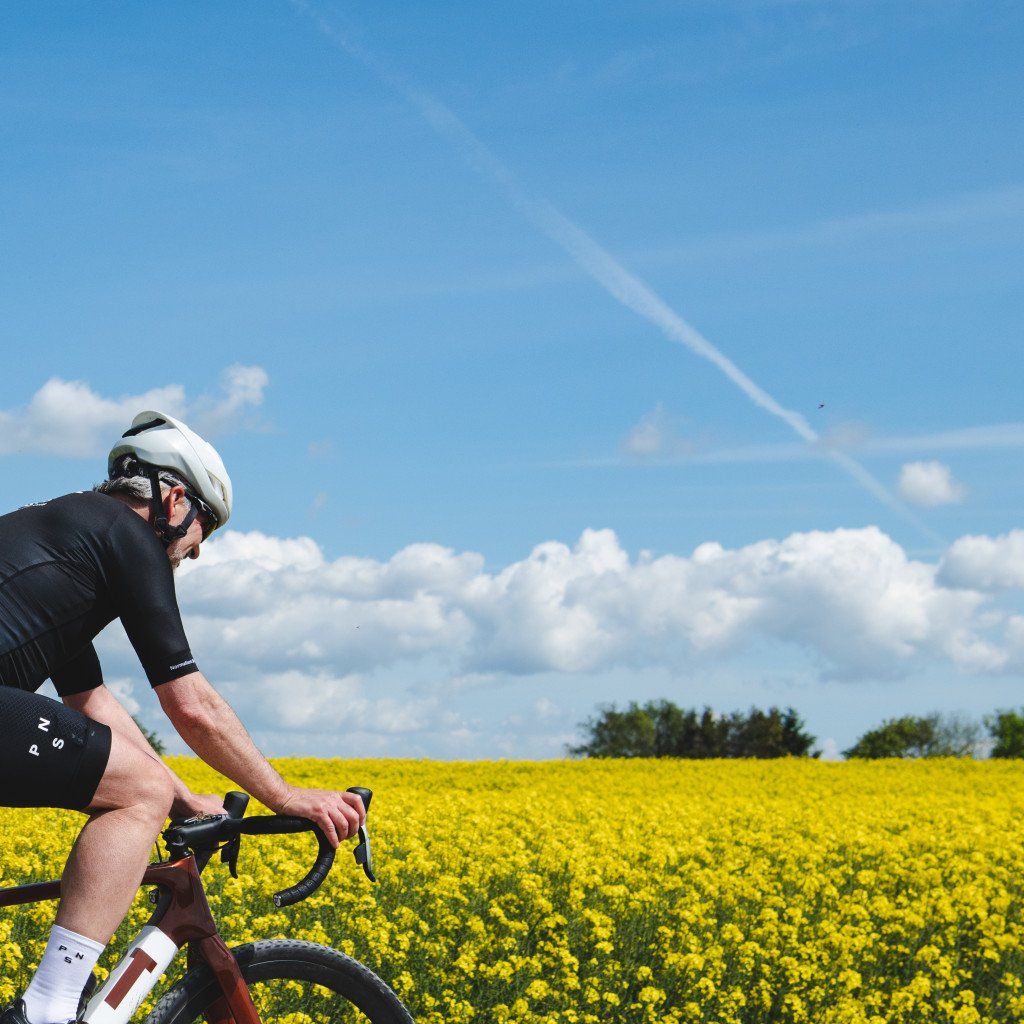 Model wearing black cycling jersey and shorts on time trial bike in yellow flower field under blue sky