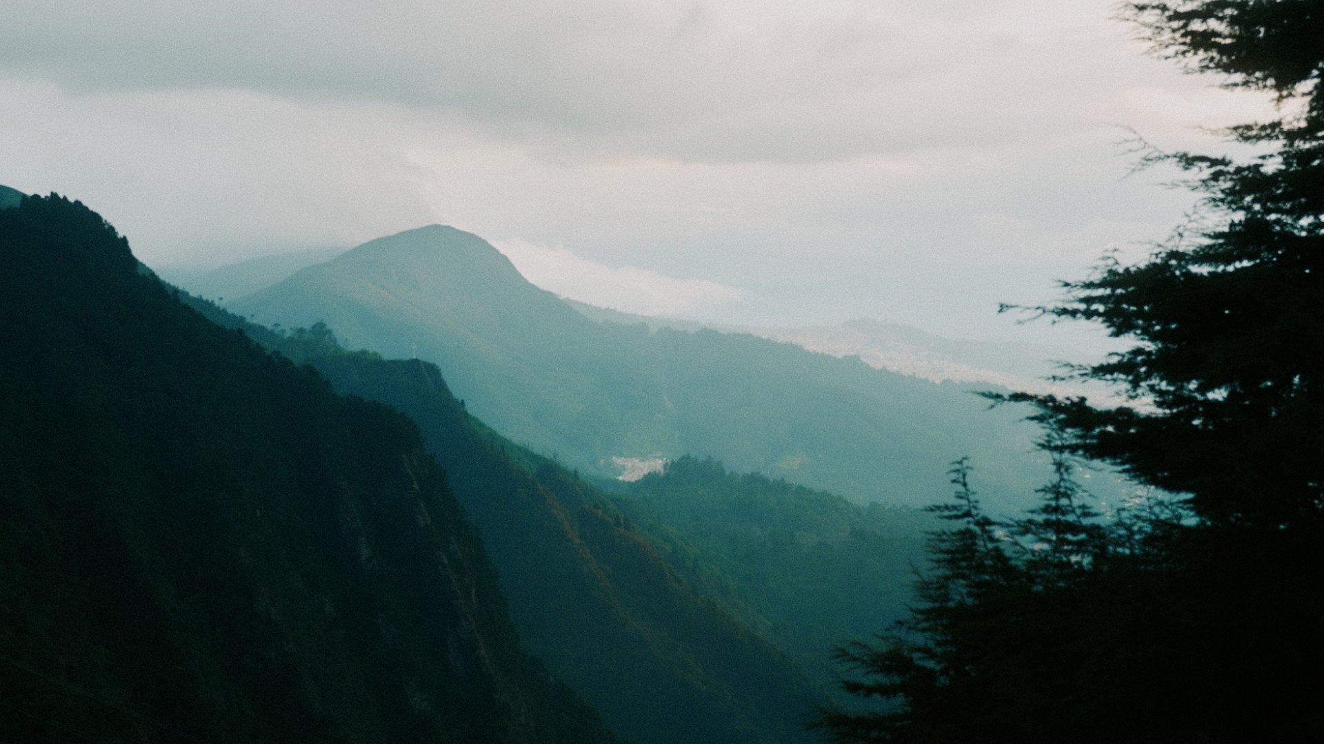 Misty mountain landscape with layered hills and forest in soft green tones under overcast sky