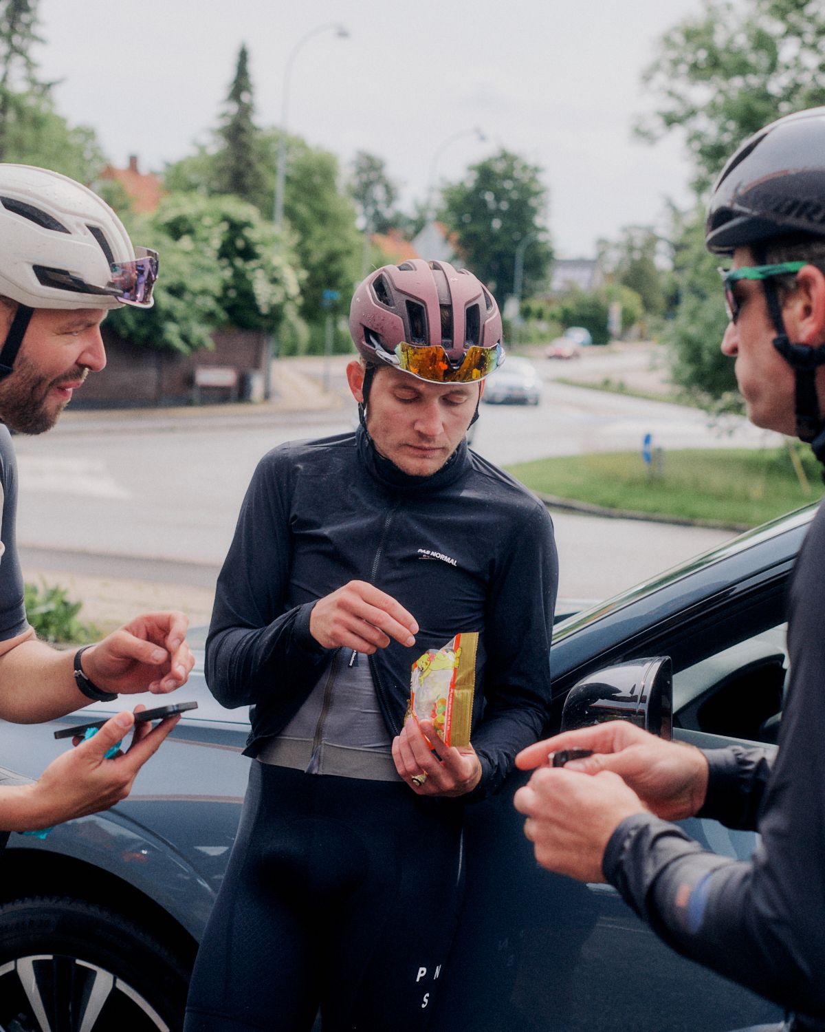 Model wearing black long-sleeve cycling jersey and bib shorts with chest branding, eating snack with teammates