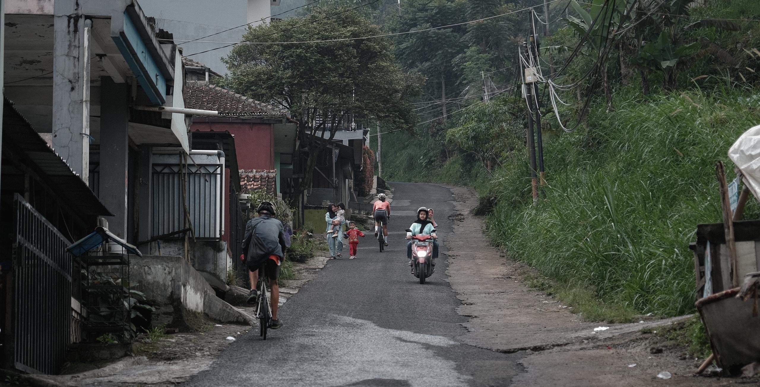 Cyclist riding through rural village street with local residents and lush green vegetation